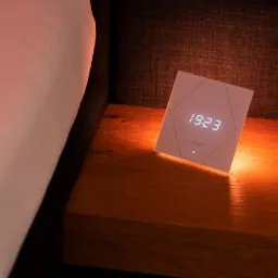 Panel sterowania inteligentnego domu Loxone, wyświetlający godzinę 19:23, umieszczony na drewnianej szafce nocnej obok łóżka z białą pościelą.