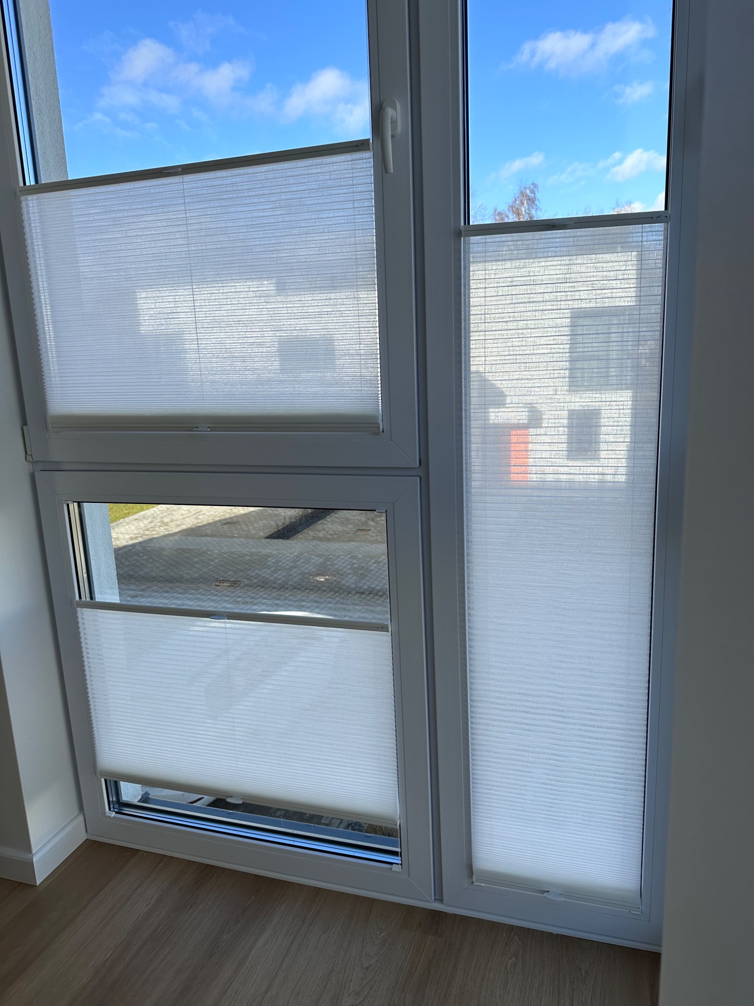 Dwu-częściowe okno z białymi, plisowanymi osłonami przeciwsłonecznymi, częściowo zasłoniętymi, widok na błękitne niebo i budynek z zewnątrz.