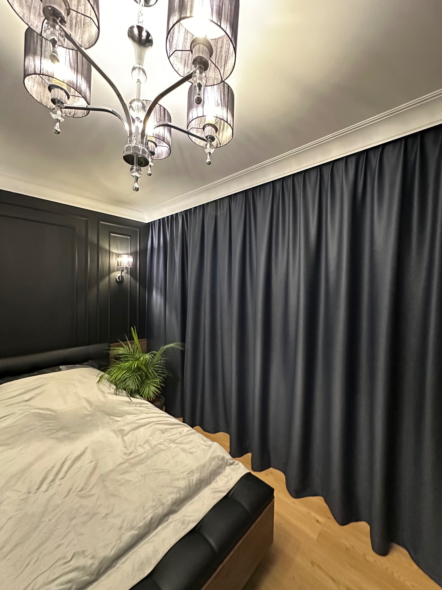 Elegancka sypialnia w ciemnej tonacji z czarnymi zasłonami, łóżkiem i dekoracyjną ścianą, oświetlona kryształowym żyrandolem i kinkietami, z rośliną doniczkową jako akcentem.