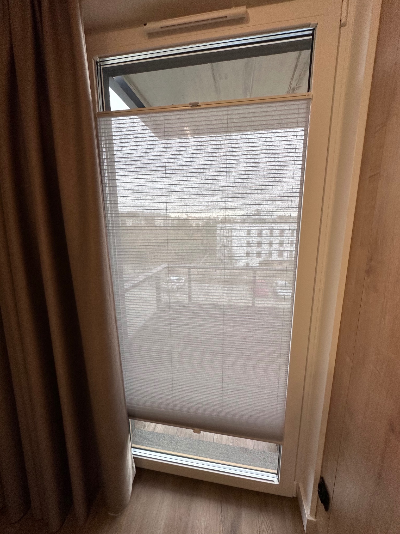 Zamknięta roleta wewnętrzna plisowana w oknie z widokiem na balkon i budynki, zasłonięta częściowo brązową zasłoną.