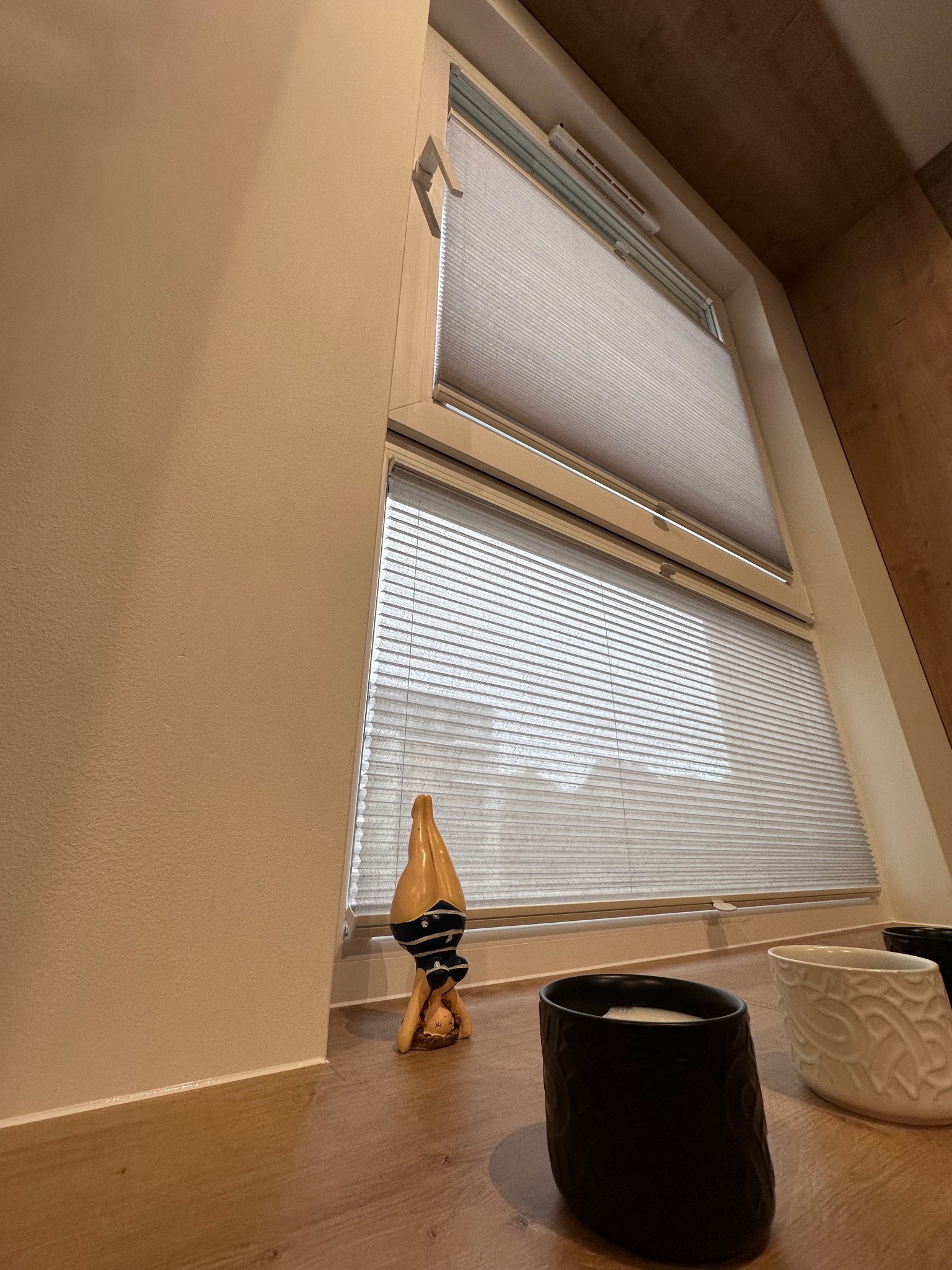 Dwuokienne okno z szarymi plisowanymi roletami, widok z dołu na fragment pokoju z dekoracyjnymi doniczkami i figurką gimnastyczki.