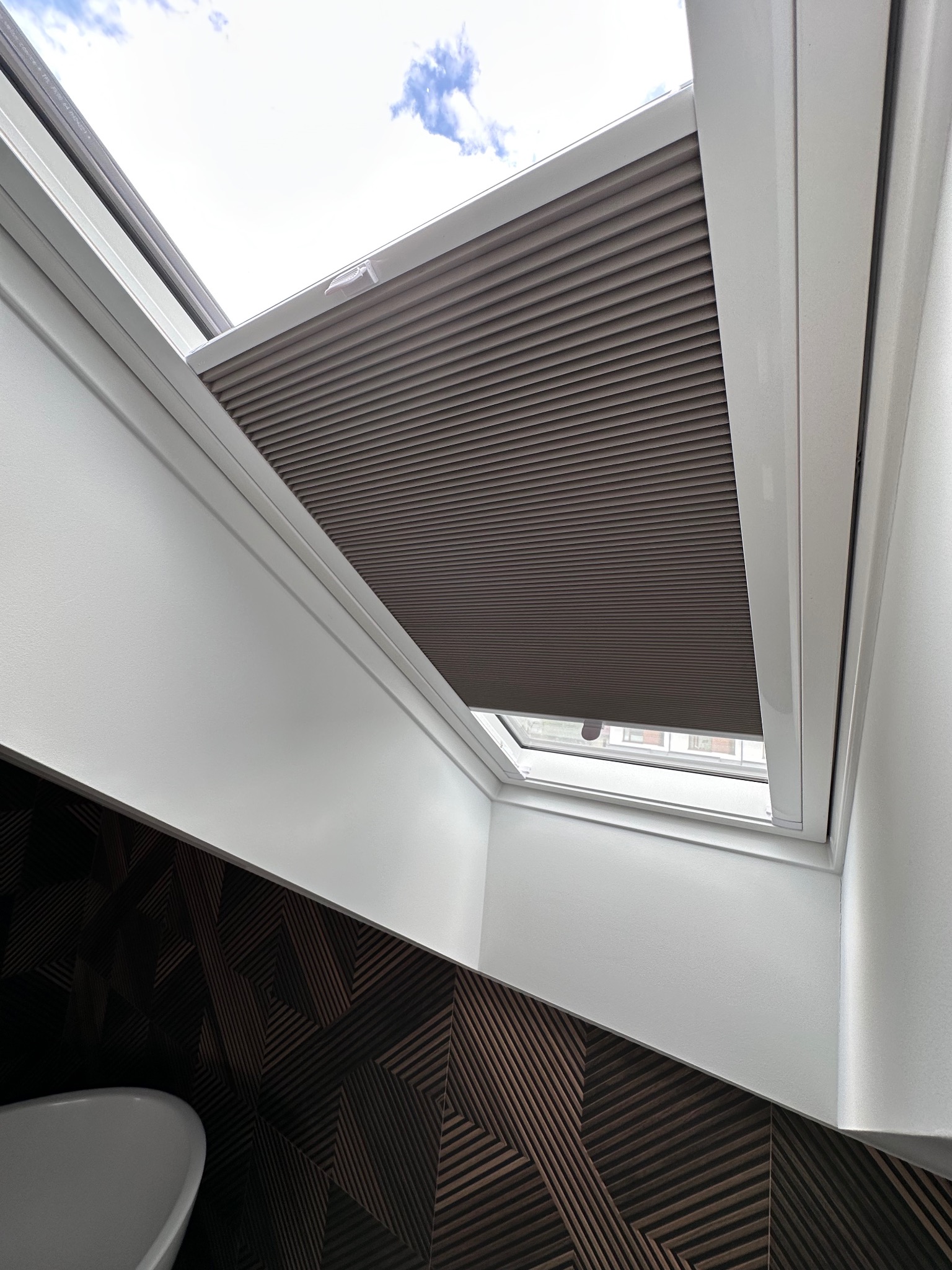 Szara, plisowana przesłona okienna w oknie dachowym, widziana od dołu, z fragmentem białej wanny i dekoracyjnej, ciemnej ściany w tle.