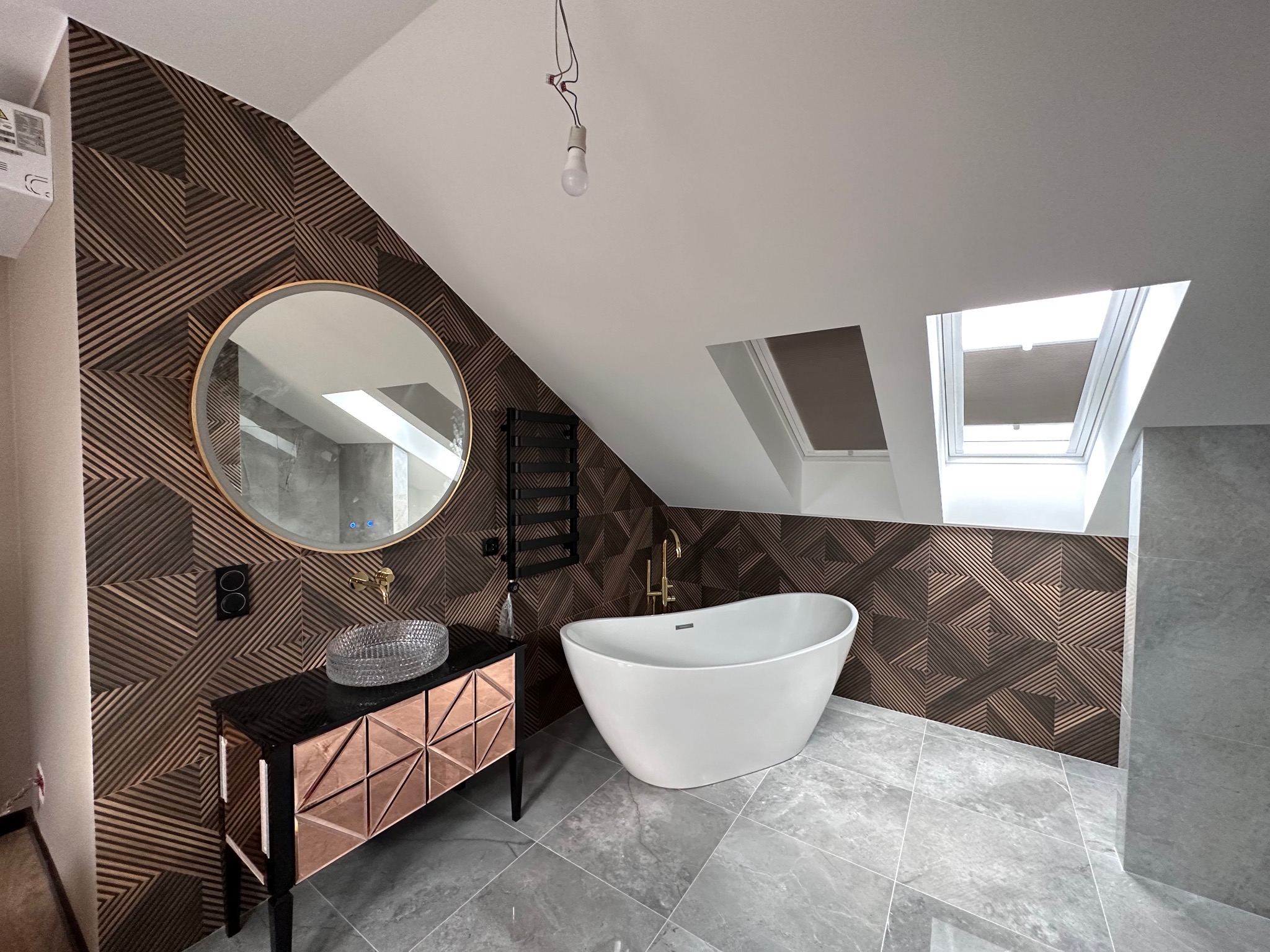 Nowoczesna łazienka na poddaszu z wolnostojącą wanną, umywalką na szafce z geometrycznym wzorem i okrągłym lustrem, wykończona panelami ściennymi w geometryczny wzór i szarą płytką podłogową.