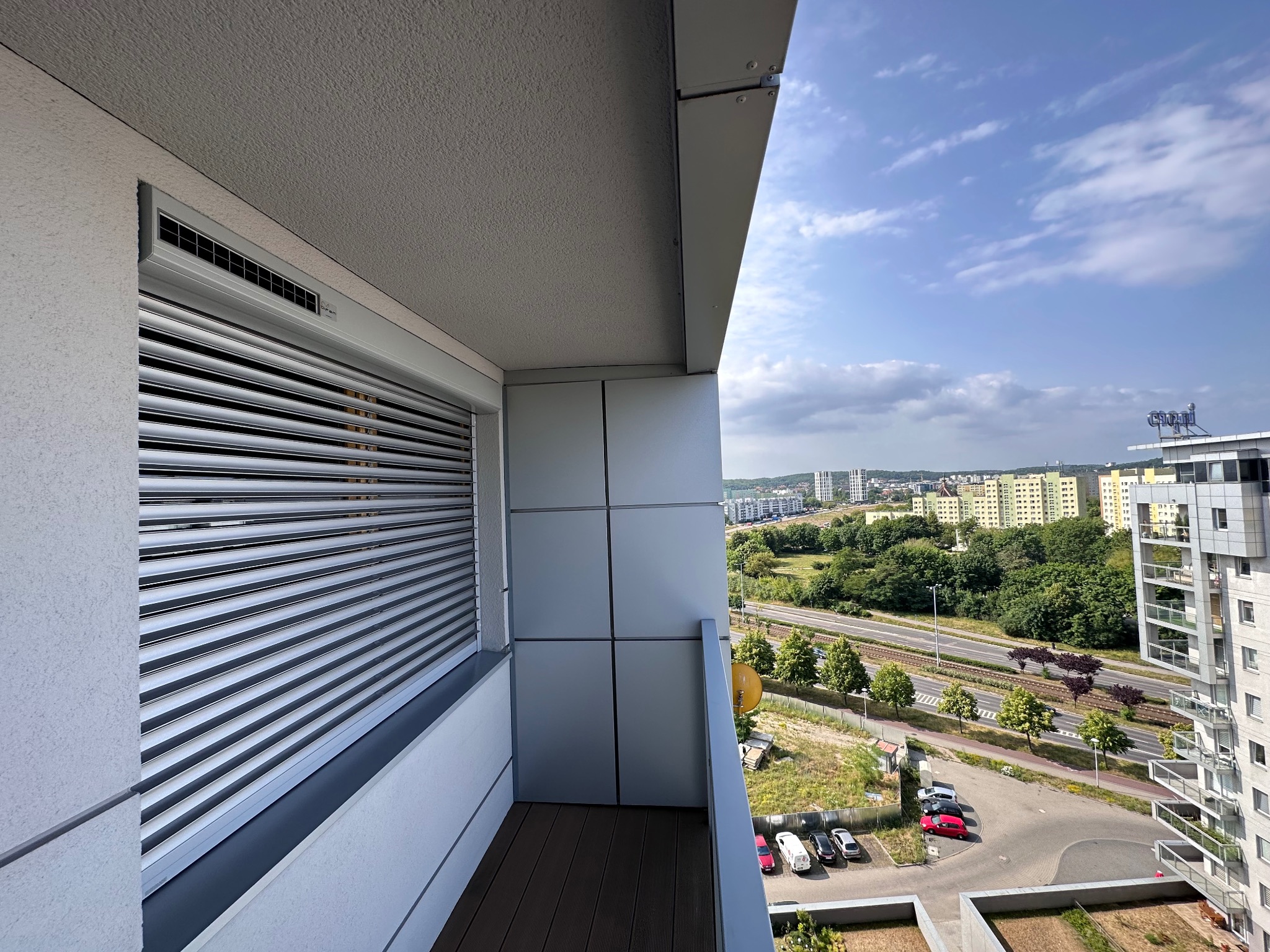 Widok z balkonu z zamontowanymi zewnętrznymi roletami aluminiowymi, na tle miejskiego krajobrazu z zielenią i budynkami.