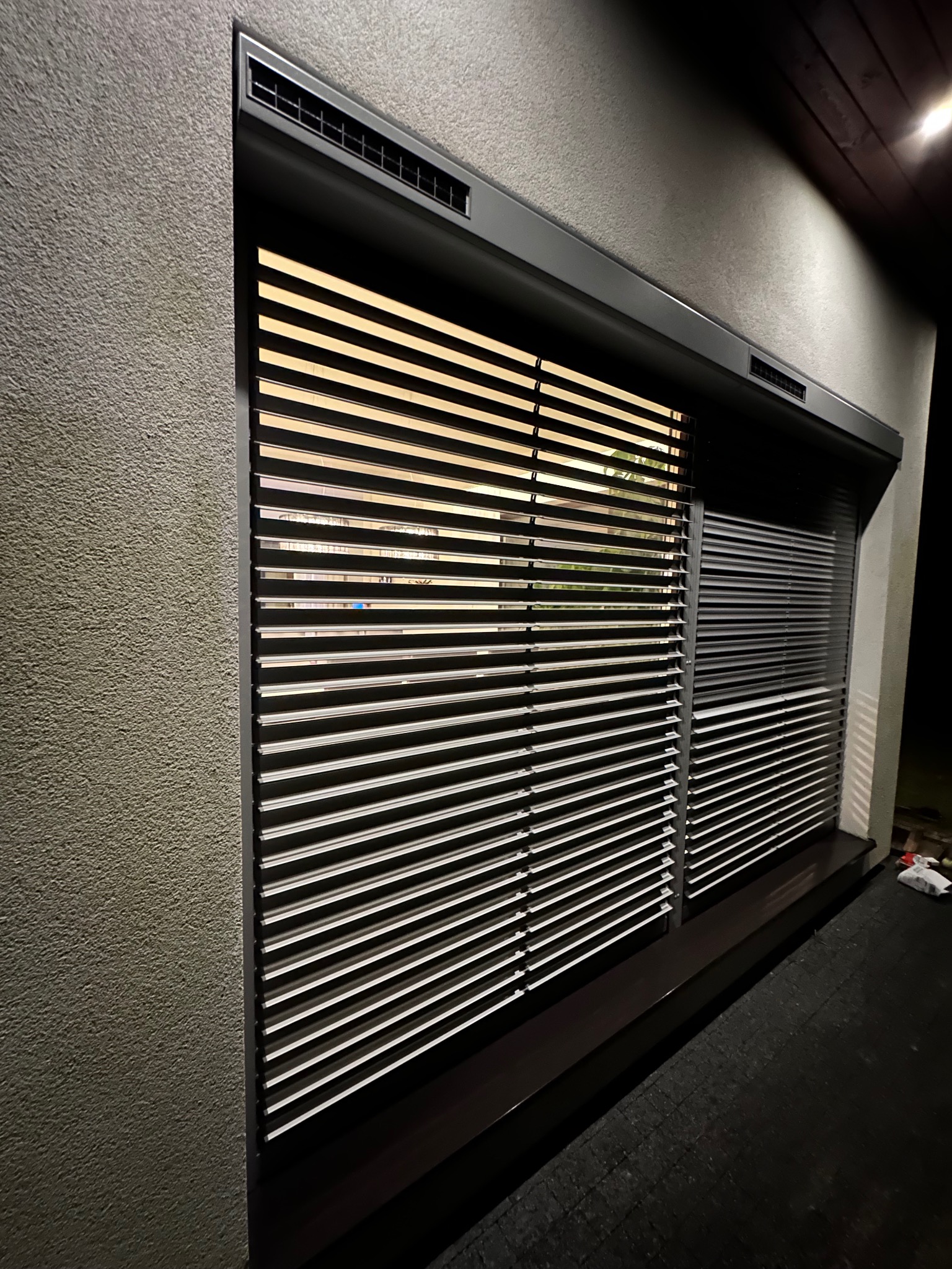 Dwa okna z szarymi, poziomymi żaluzjami zewnętrznymi, jedno częściowo otwarte, drugie zamknięte, widoczne na tle szarej, teksturowanej ściany budynku, oświetlone z góry.