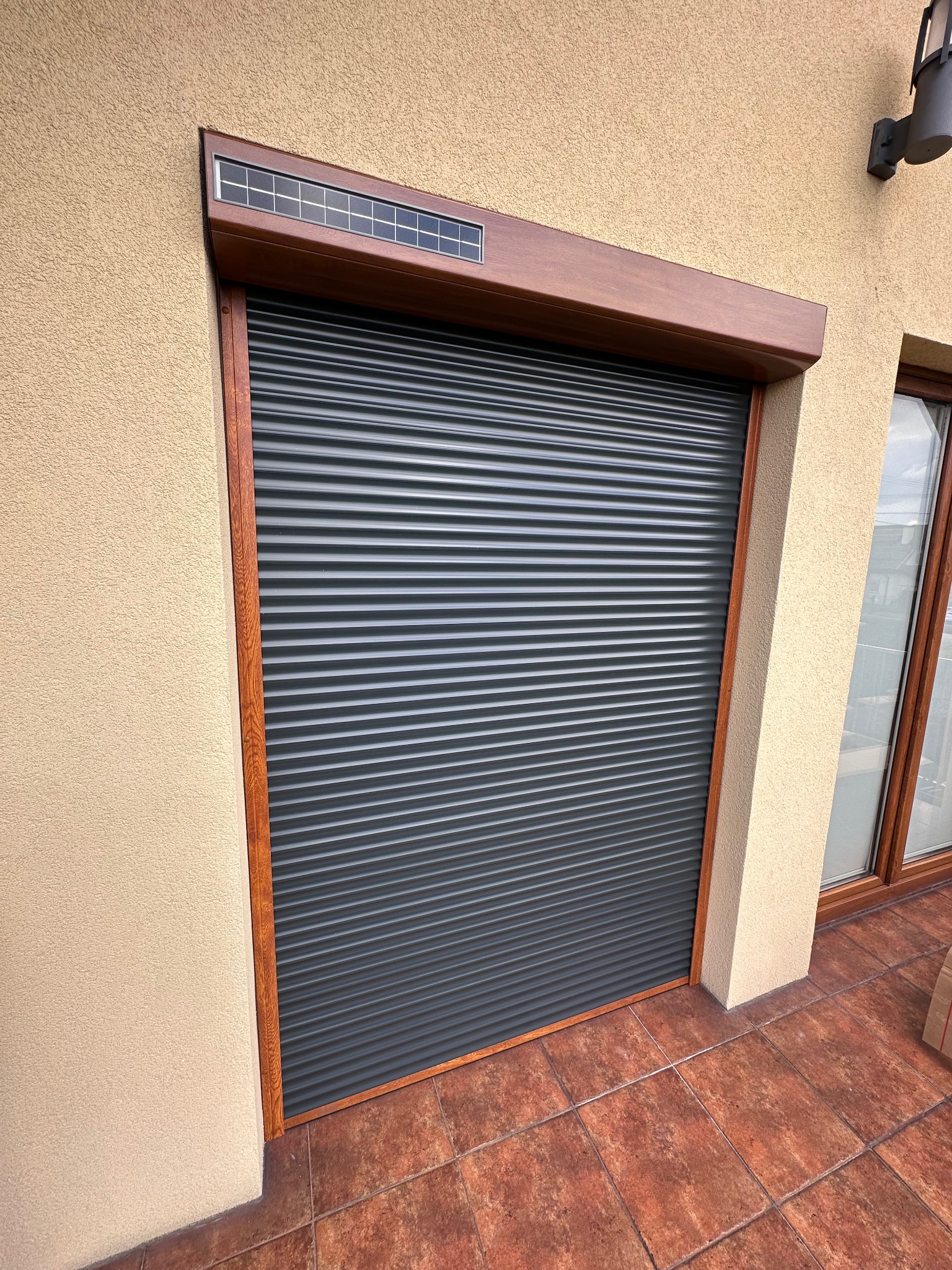 Zamknięta, grafitowa roleta zewnętrzna z brązową ramą i panelem solarnym u góry, zamontowana na elewacji budynku z terakotową podłogą.