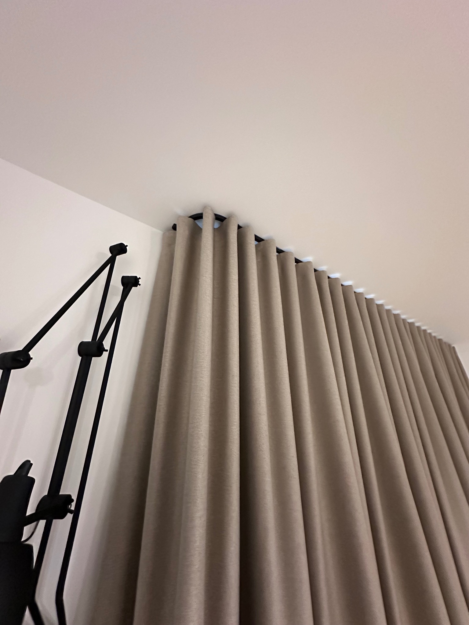 Beżowe zasłony z równomiernymi fałdami zawieszone na czarnym karniszu, widoczne z dołu pod kątem, obok czarna lampa podłogowa.