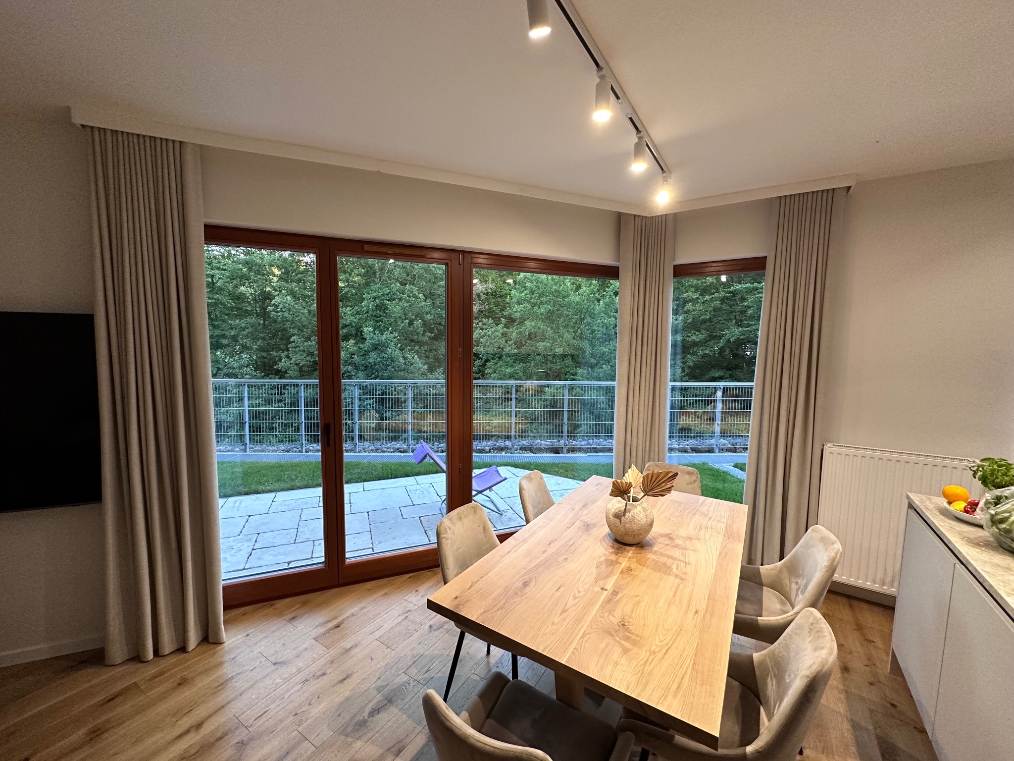 Jasne, przestronne wnętrze jadalni z dużym oknem tarasowym, beżowymi zasłonami po bokach, drewnianym stołem, krzesłami obitymi welurem i minimalistycznym oświetleniem punktowym na suficie.