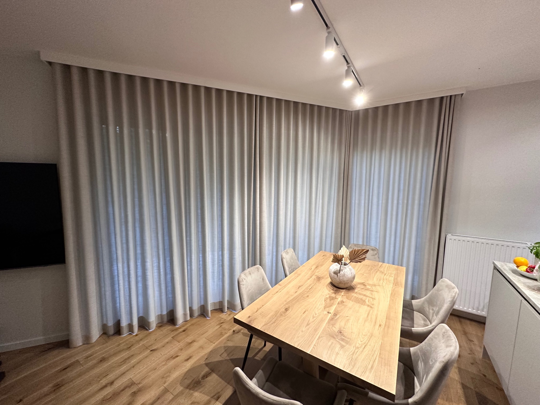 Beżowe, marszczone zasłony do podłogi w nowoczesnym salonie z drewnianym stołem i krzesłami w odcieniach szarości, oświetlone reflektorami sufitowymi.