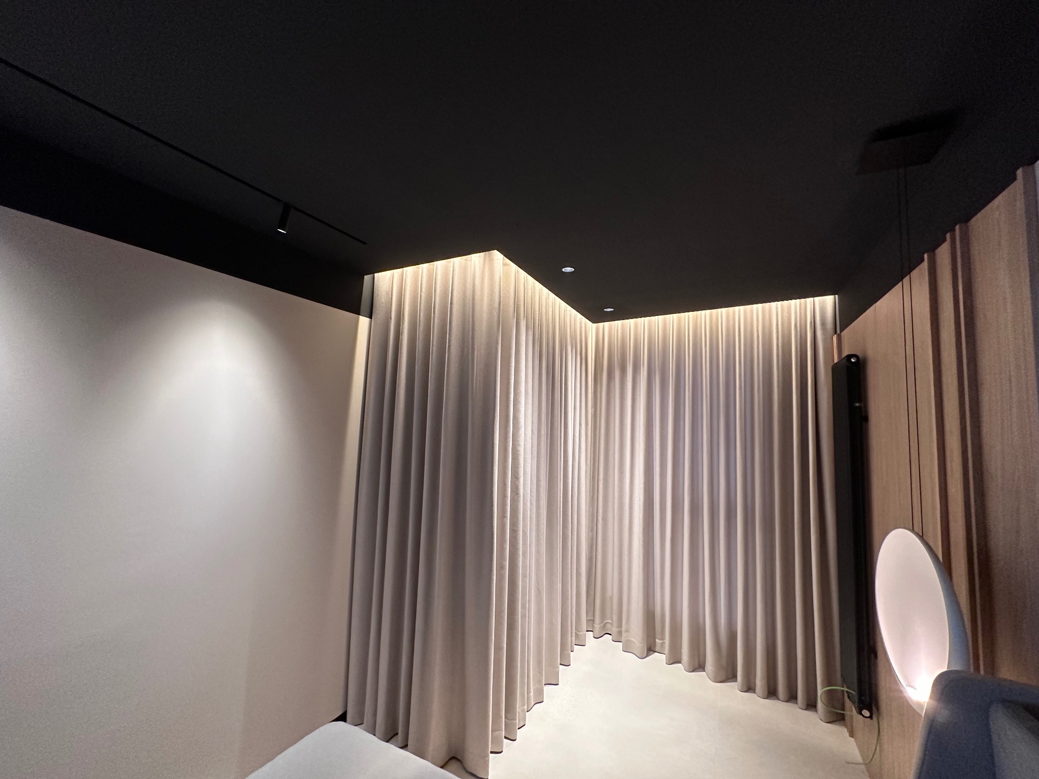 Eleganckie, jasne zasłony zakrywające całą ścianę, podświetlone od góry, w minimalistycznym wnętrzu z czarnym sufitem i nowoczesnym oświetleniem punktowym.