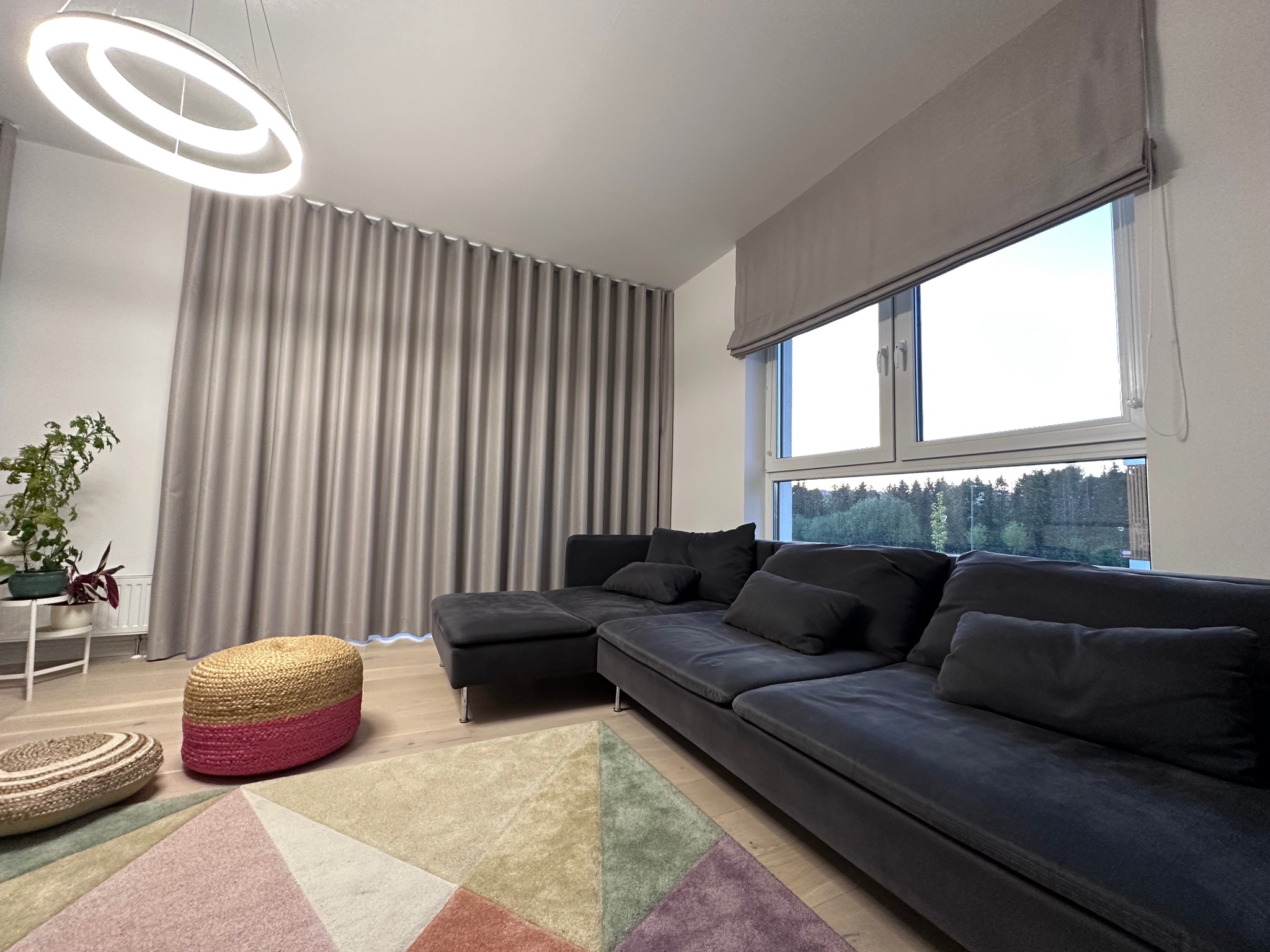 Nowoczesny salon z szarą, falującą zasłoną, szarą roletą rzymską, ciemną sofą modułową i geometrycznym dywanem w pastelowych kolorach.