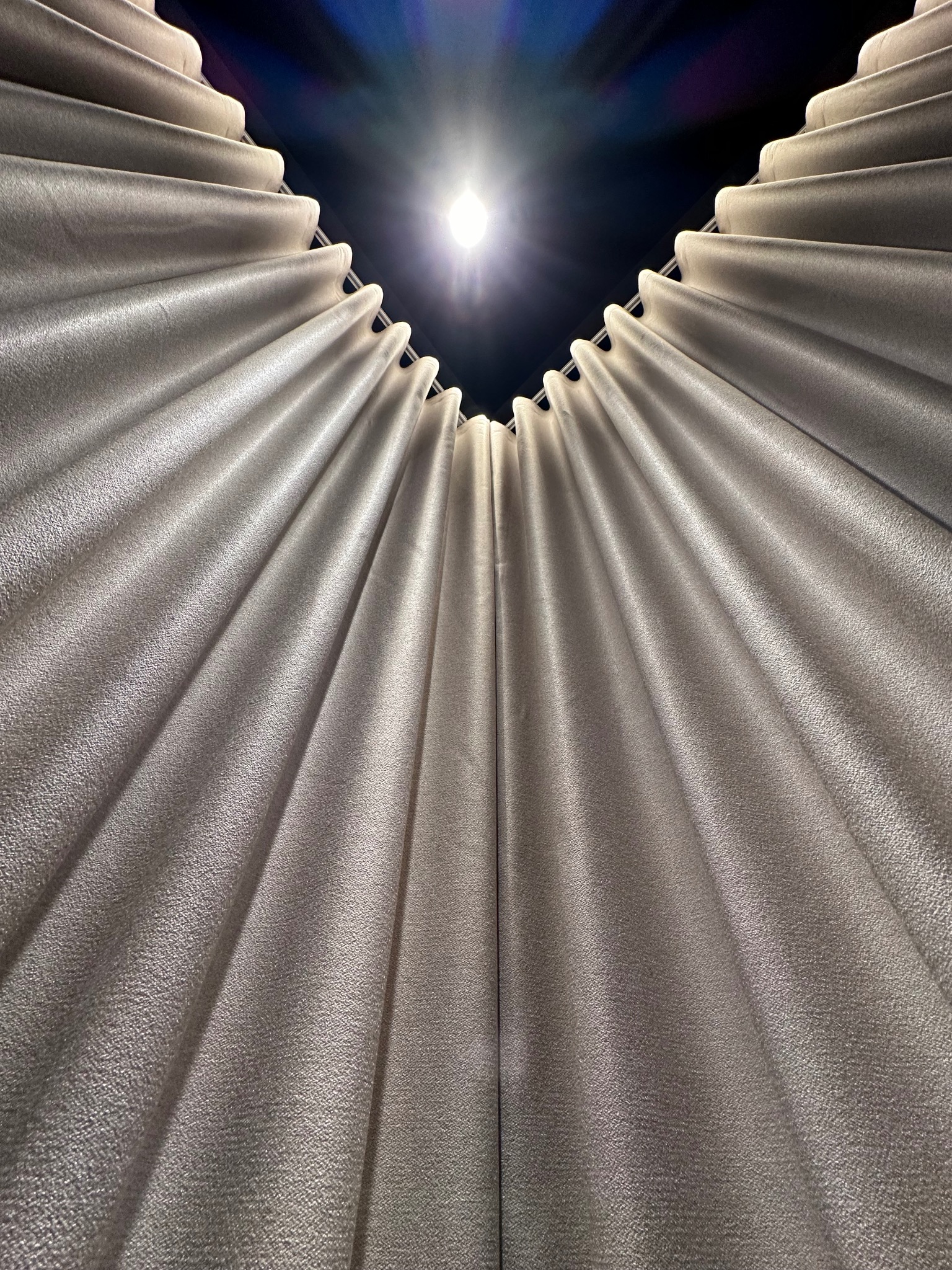 Widok z dołu na falujące, beżowe zasłony zawieszone na szynie, z jasnym światłem w tle.