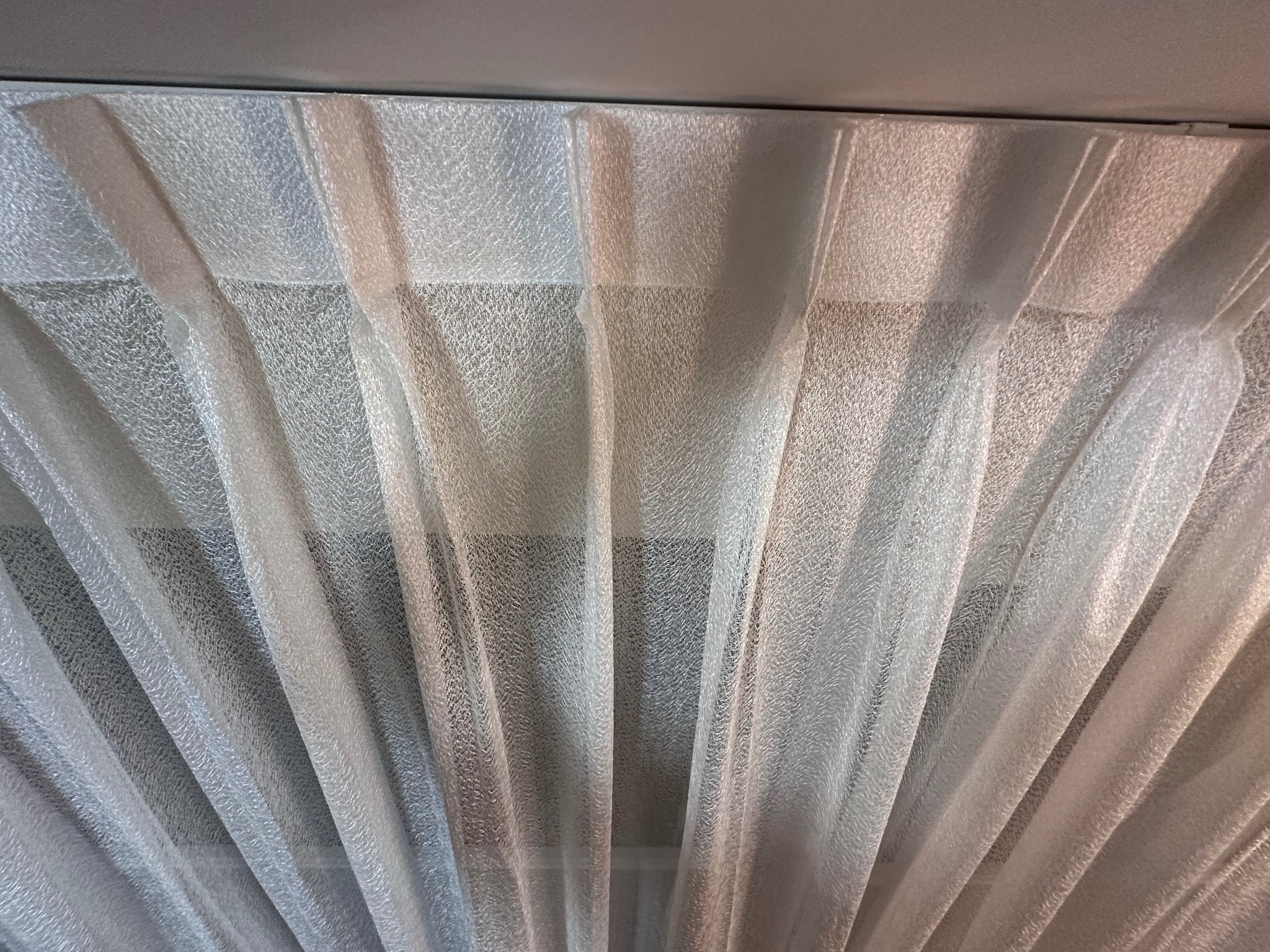 Delikatna, biała, półprzezroczysta tkanina zasłaniająca okno, ułożona w regularne fałdy, widok od dołu, z widocznym karniszem.