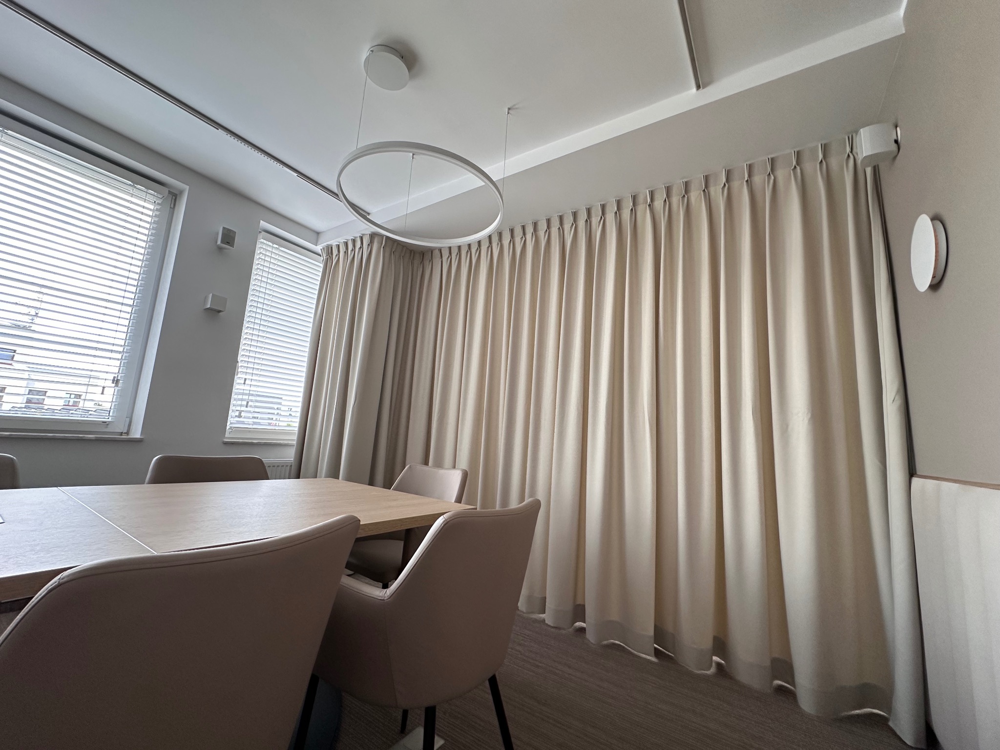 Nowoczesna sala konferencyjna z beżowymi zasłonami zakrywającymi całą ścianę, minimalistycznym okrągłym żyrandolem i dużym stołem otoczonym krzesłami w kolorze nude.