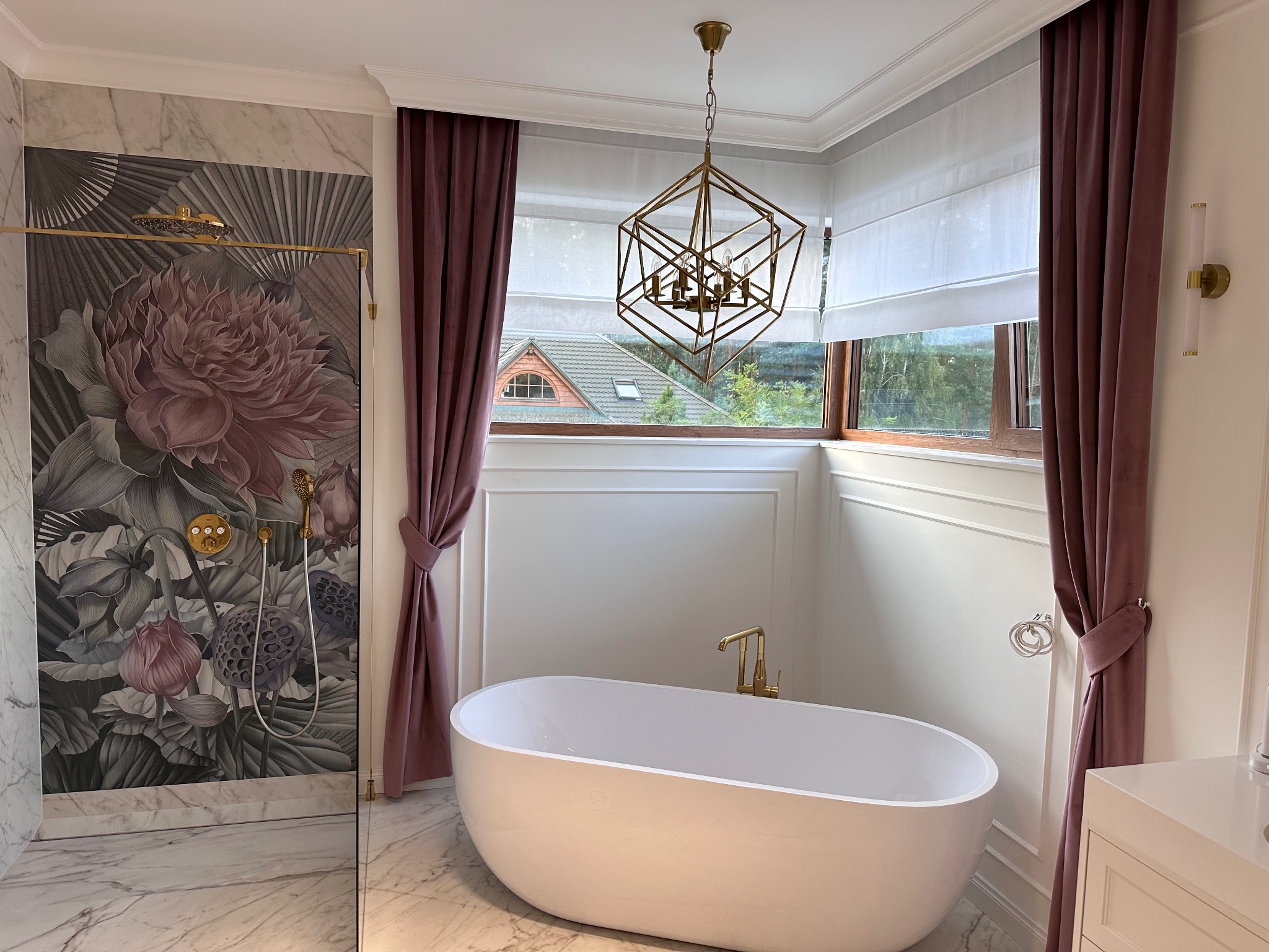Elegancka łazienka z wolnostojącą wanną, dekoracyjną tapetą z motywem kwiatowym, nowoczesną lampą wiszącą i bordowymi zasłonami oraz roletami rzymskimi w oknie.
