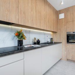 Marta Rudnicka (Rudnicka Interiors) - Nowoczesna kuchnia z drewnianymi szafkami, białymi frontami dolnymi, czarnym blatem i marmurowym backsplash'em, ozdobiona kwiatami w czarnym wazonie i dekoracyjnymi wazonami.