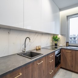 Marta Rudnicka (Rudnicka Interiors) - Nowoczesna kuchnia z białymi szafkami górnymi, drewnianymi szafkami dolnymi z złotymi uchwytami, marmurowym backsplashiem, czarnym zlewem i czarną baterią, oświetleniem LED pod szafkami i roślinami...