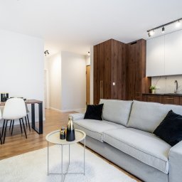 Marta Rudnicka (Rudnicka Interiors) - Jasne, przestronne wnętrze mieszkania z szarą sofą, okrągłym stolikiem kawowym z dekoracyjnymi flakonami, stołem jadalnym z białymi krzesłami i aneksem kuchennym z drewnianymi i białymi szafkami.