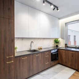Marta Rudnicka (Rudnicka Interiors) - Nowoczesna kuchnia z białymi szafkami górnymi i ciemnobrązowymi dolnymi, marmurowy backsplash, czarny zlew i bateria, złote uchwyty, oświetlenie punktowe