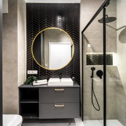 Marta Rudnicka (Rudnicka Interiors) - Nowoczesna łazienka z czarną armaturą, okrągłym lustrem w złotej ramie i płytkami ułożonymi w jodełkę na ścianie za umywalką.
