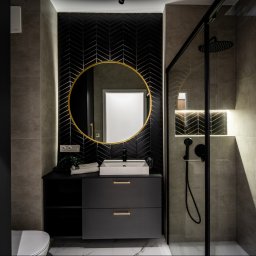 Marta Rudnicka (Rudnicka Interiors) - Nowoczesna łazienka z czarną armaturą, okrągłym lustrem w złotej ramie i płytkami ułożonymi w jodełkę na ścianie za umywalką. Kabina prysznicowa ze szklaną ścianą.