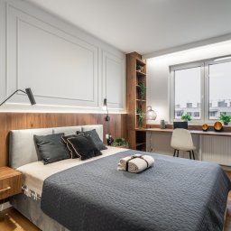 Marta Rudnicka (Rudnicka Interiors) - Nowoczesna sypialnia z drewnianym panelem za łóżkiem, minimalistycznymi lampami ściennymi, biurkiem przy oknie z roślinami doniczkowymi i dekoracjami, oraz stolikiem nocnym z ozdobną butelką.