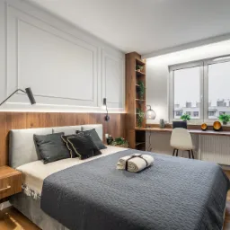 Nowoczesna sypialnia z drewnianym panelem za łóżkiem, minimalistycznymi lampami ściennymi, biurkiem przy oknie z roślinami doniczkowymi i dekoracjami, oraz stolikiem nocnym z ozdobną butelką.