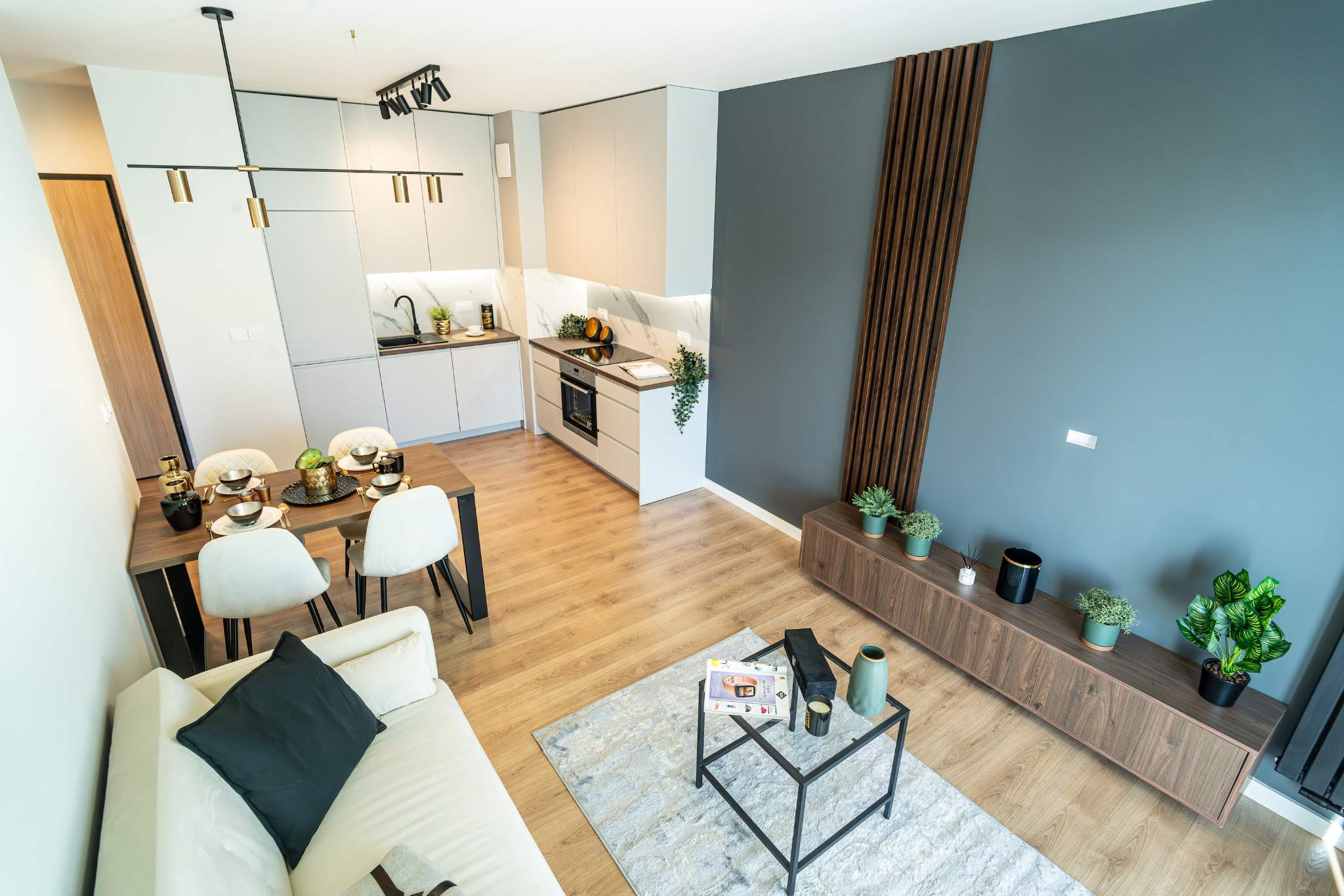 Widok z góry na nowocześnie urządzone mieszkanie z aneksem kuchennym, drewnianym stołem jadalnym, białymi krzesłami, szarą ścianą z dekoracyjnymi drewnianymi listwami i niską szafką RTV z roślinami...