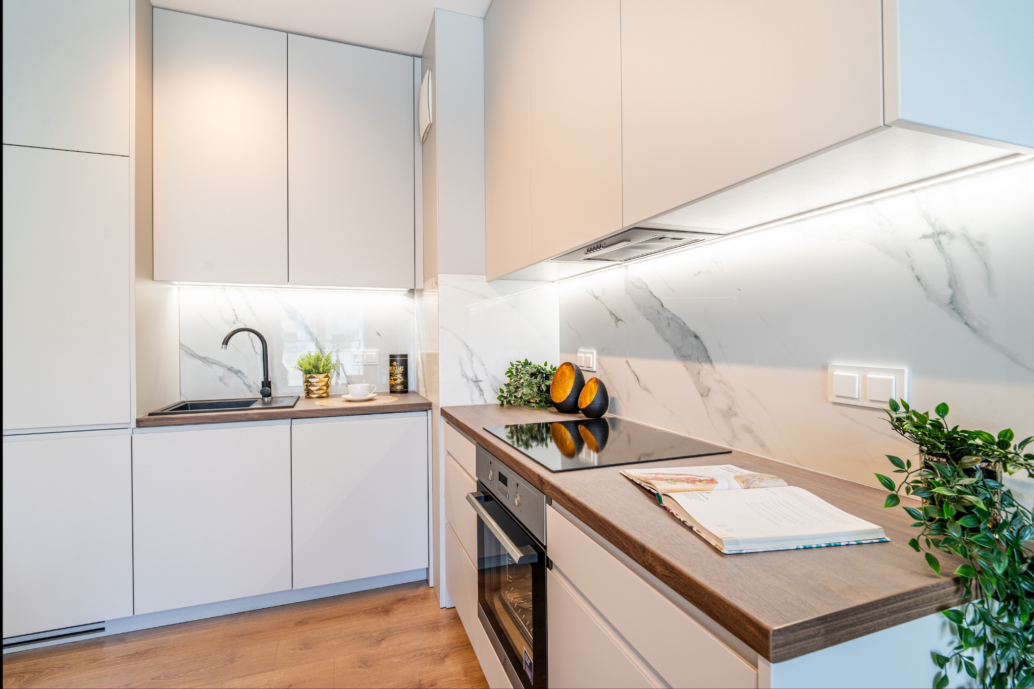 Jasna kuchnia z białymi szafkami, marmurowym backsplashiem i drewnianym blatem, na którym leży otwarta książka kucharska i stoi roślina doniczkowa.