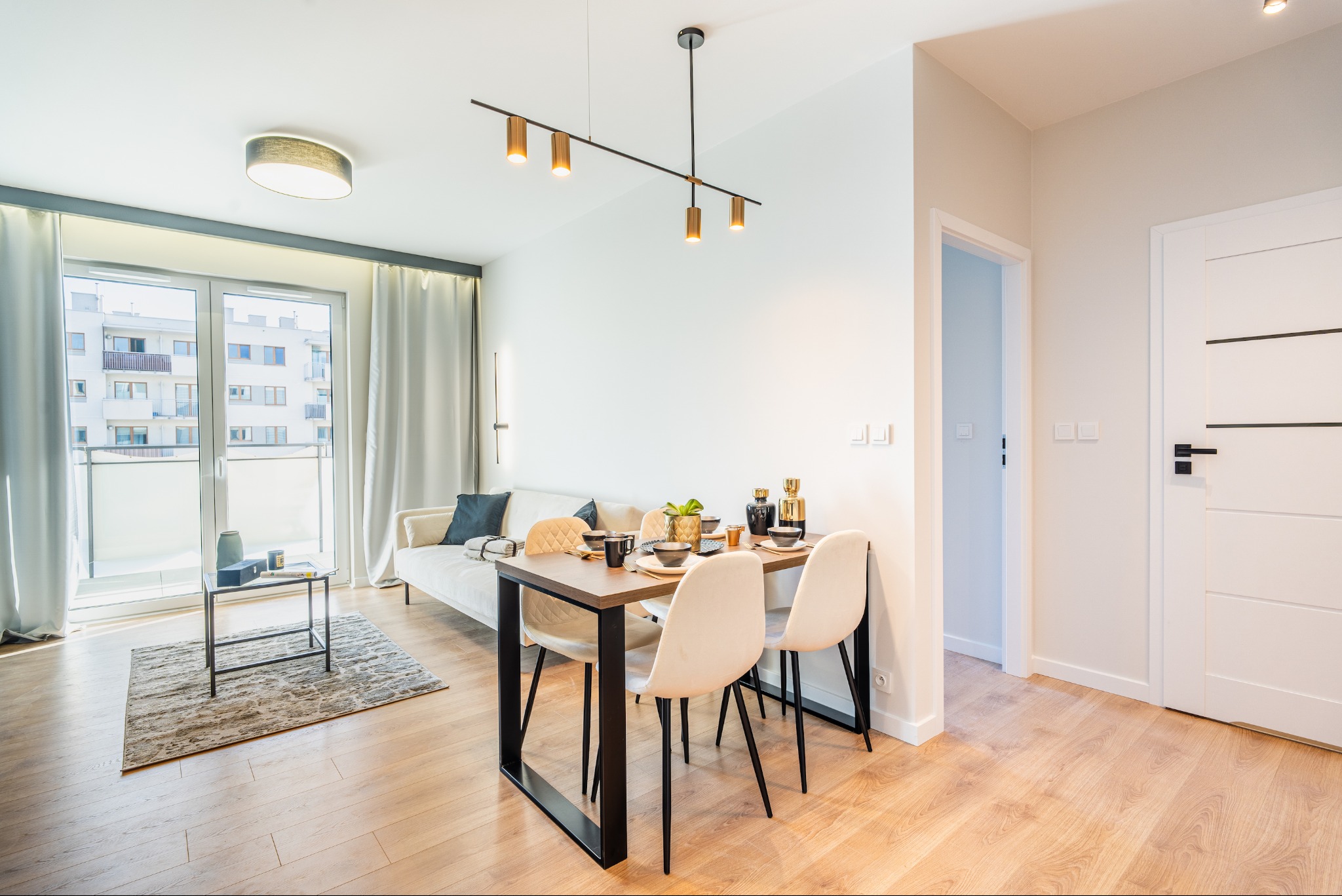 Jasne wnętrze mieszkania z drewnianą podłogą, stołem jadalnym z nakryciem, kremowymi krzesłami, sofą i balkonem z widokiem na budynek mieszkalny