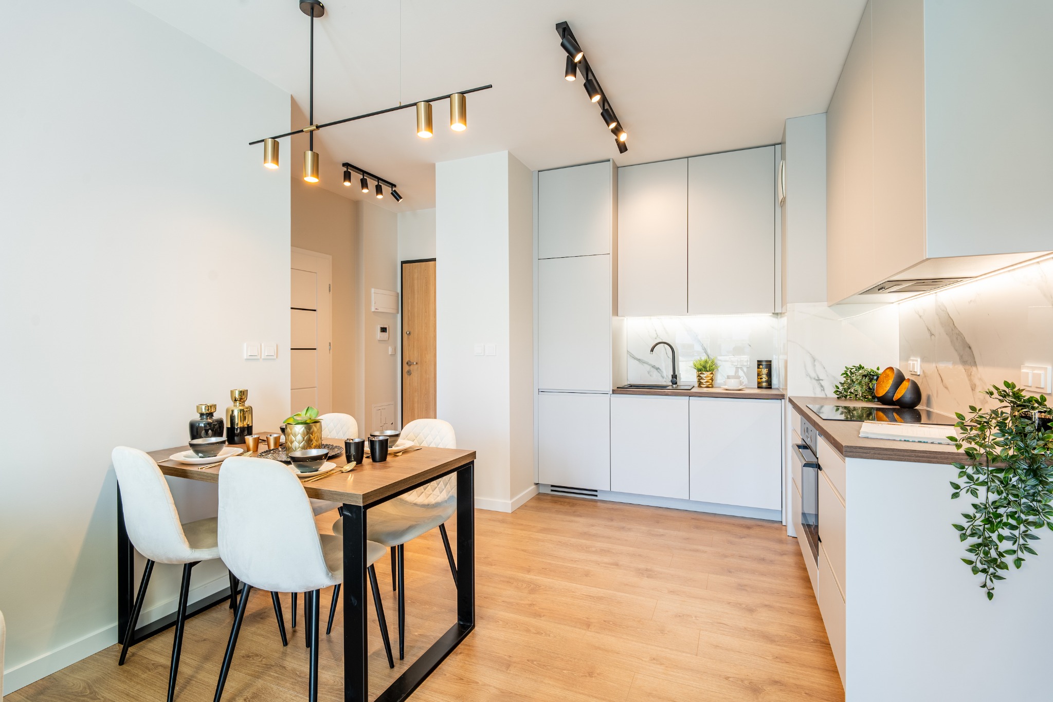 Jasne, nowoczesne wnętrze: stół jadalny z białymi krzesłami, minimalistyczna kuchnia z białymi szafkami i marmurowym backsplashiem, oświetlenie punktowe na suficie.