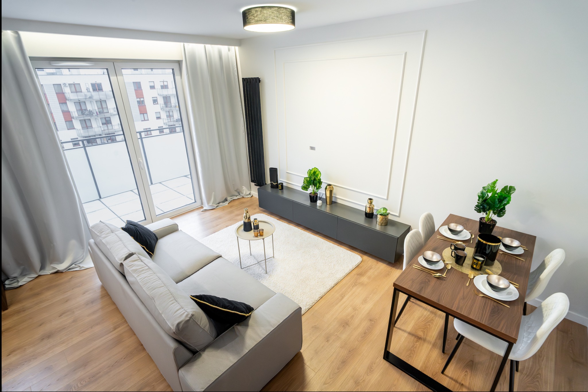 Jasne, nowocześnie urządzone wnętrze mieszkania z balkonem, drewnianą podłogą, szarą sofą z czarnymi poduszkami, stołem jadalnym z czterema krzesłami i dekoracjami, szarą szafką RTV z roślinami...