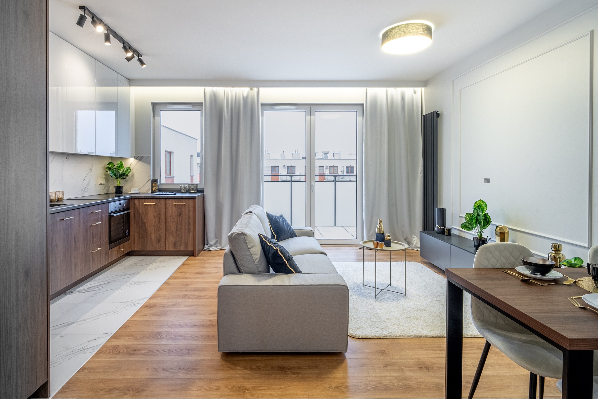 Nowoczesne wnętrze mieszkania z aneksem kuchennym, szarą sofą z poduszkami, drewnianym stołem jadalnym i białym dywanem shaggy, oświetlone punktowo i lampą sufitową.