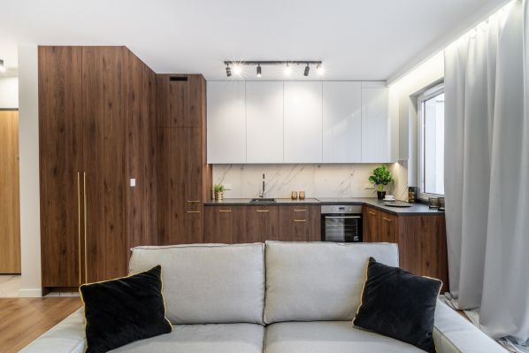 Nowoczesne wnętrze mieszkania z otwartą kuchnią, widok z salonu na zabudowę kuchenną z białymi szafkami górnymi i drewnopodobnymi dolnymi, marmurowy backsplash, oświetlenie punktowe i sofa...
