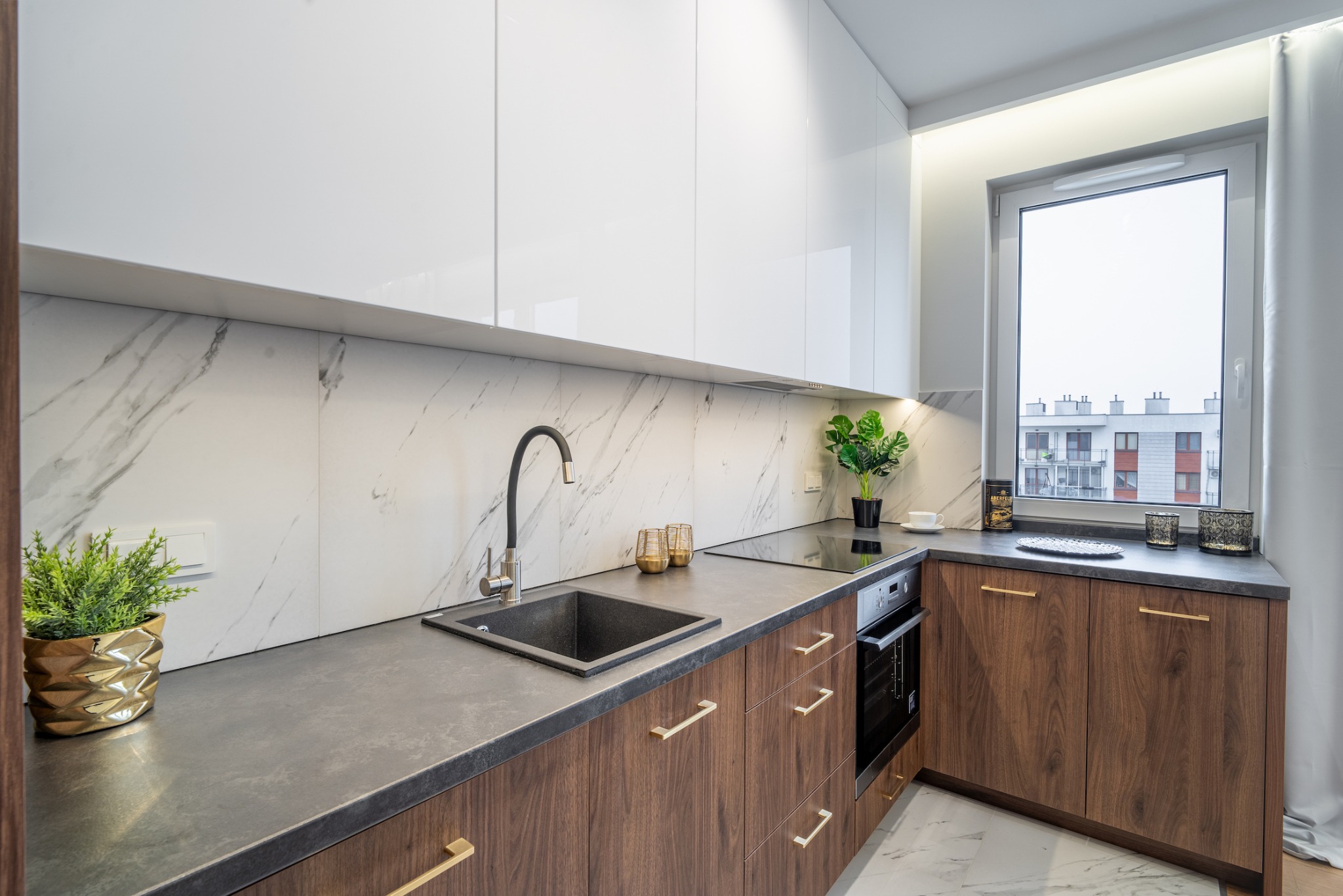 Nowoczesna kuchnia z białymi szafkami górnymi, drewnianymi szafkami dolnymi z złotymi uchwytami, marmurowym backsplashiem, czarnym zlewem i czarną baterią, oświetleniem LED pod szafkami i roślinami...