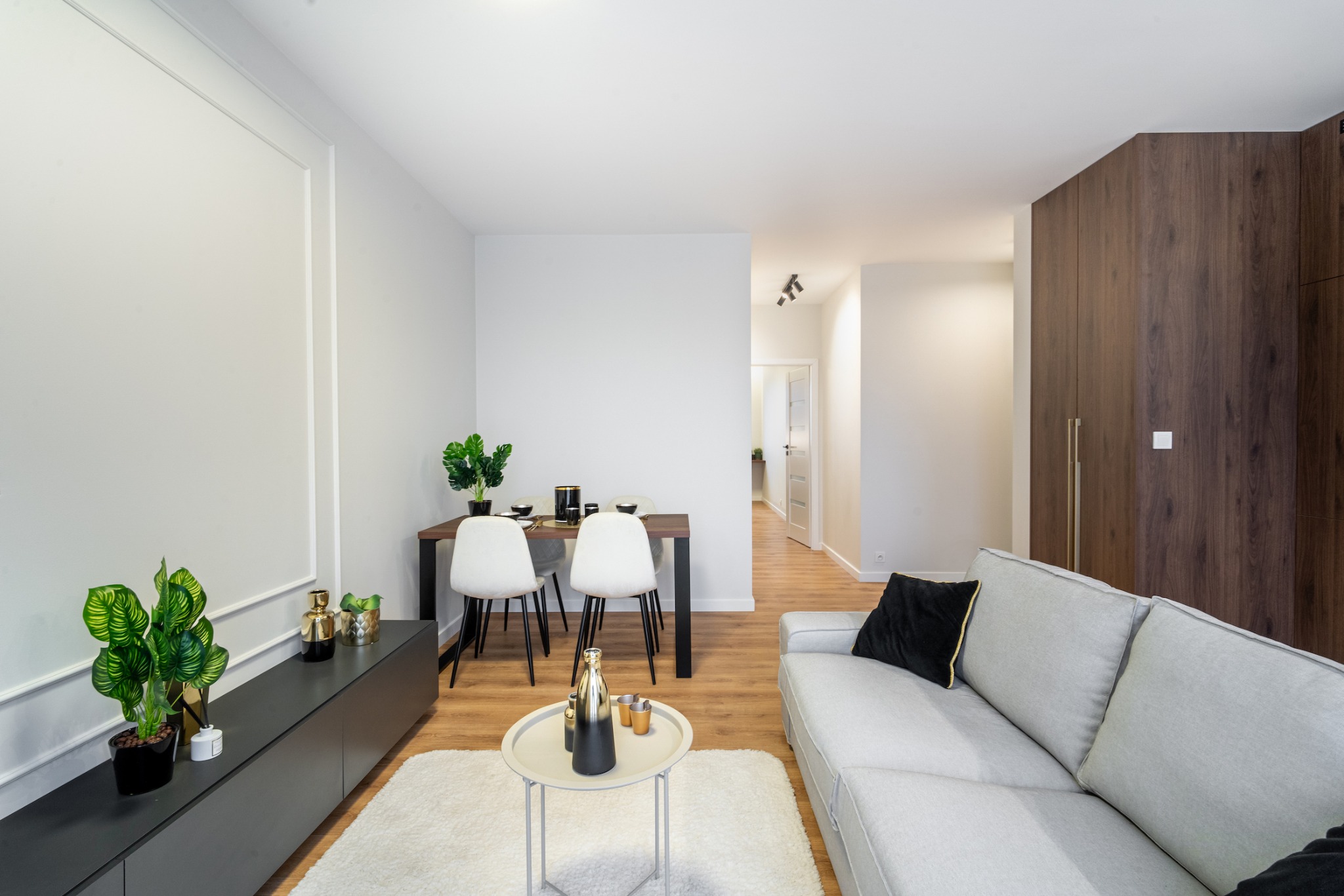 Jasne wnętrze salonu z jadalnią: szara sofa z czarną poduszką, stolik kawowy z dekoracjami, stół z krzesłami i roślinami w doniczkach, białe ściany z dekoracyjną listwą, drewniana podłoga i dywan...