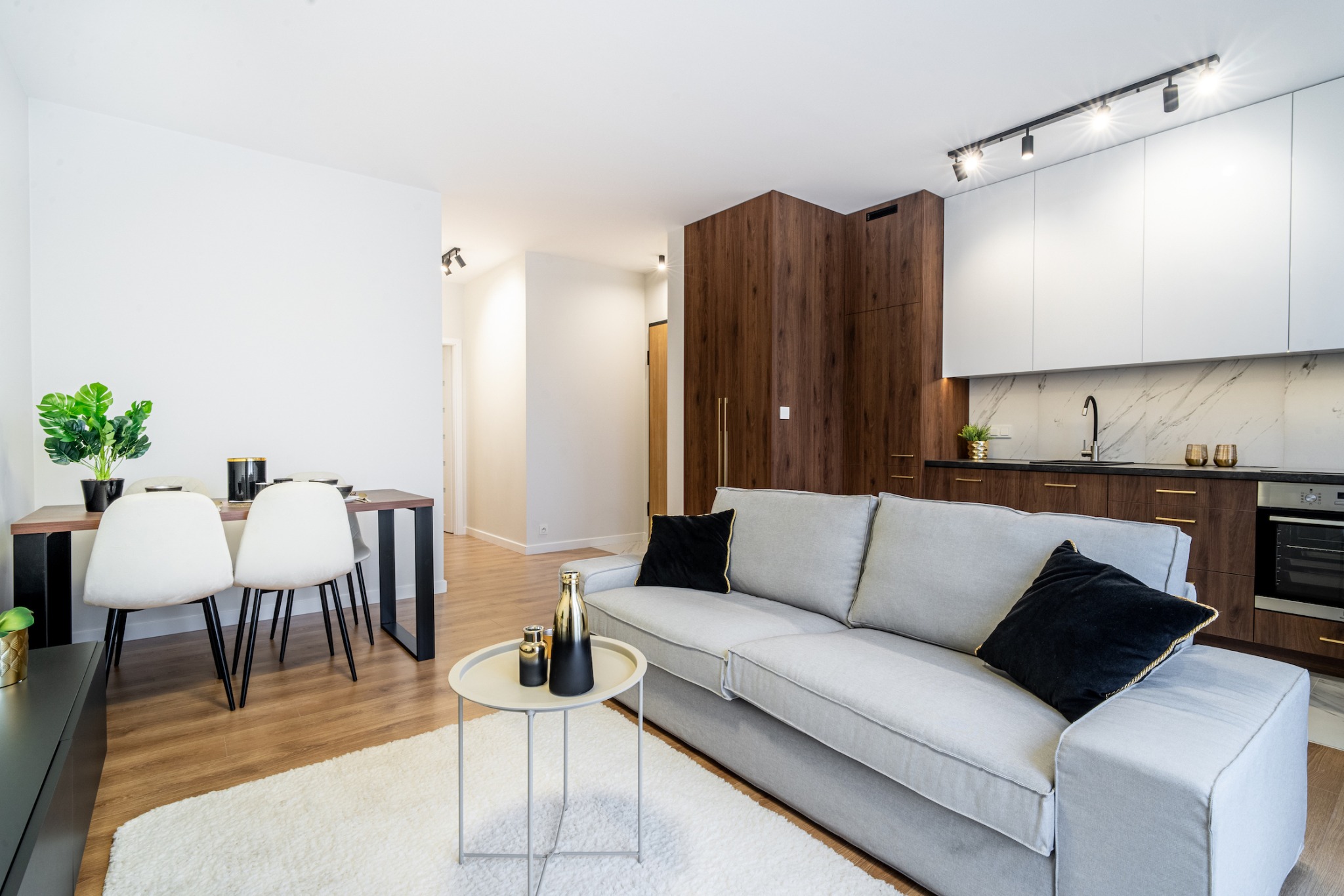 Jasne, przestronne wnętrze mieszkania z szarą sofą, okrągłym stolikiem kawowym z dekoracyjnymi flakonami, stołem jadalnym z białymi krzesłami i aneksem kuchennym z drewnianymi i białymi szafkami.