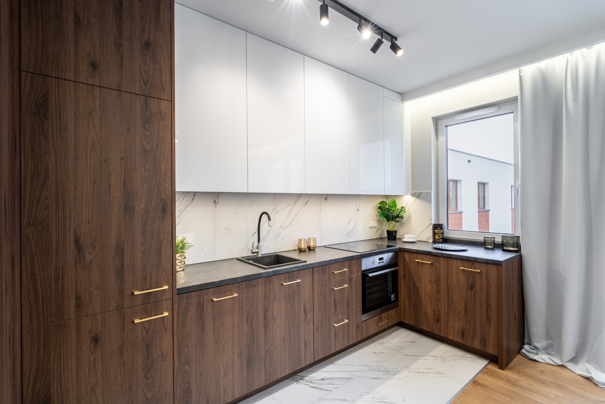 Nowoczesna kuchnia z białymi szafkami górnymi i ciemnobrązowymi dolnymi, marmurowy backsplash, czarny zlew i bateria, złote uchwyty, oświetlenie punktowe
