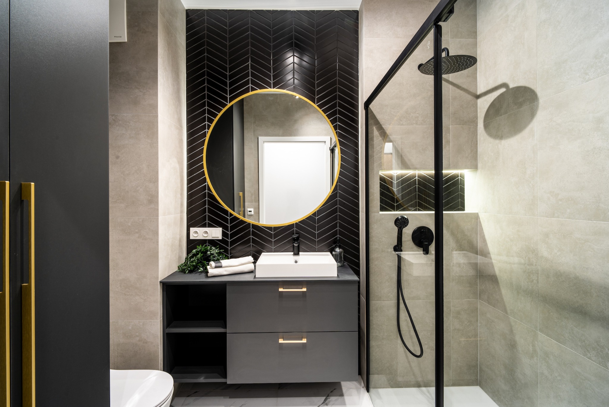 Nowoczesna łazienka z czarną armaturą, okrągłym lustrem w złotej ramie i płytkami ułożonymi w jodełkę na ścianie za umywalką.