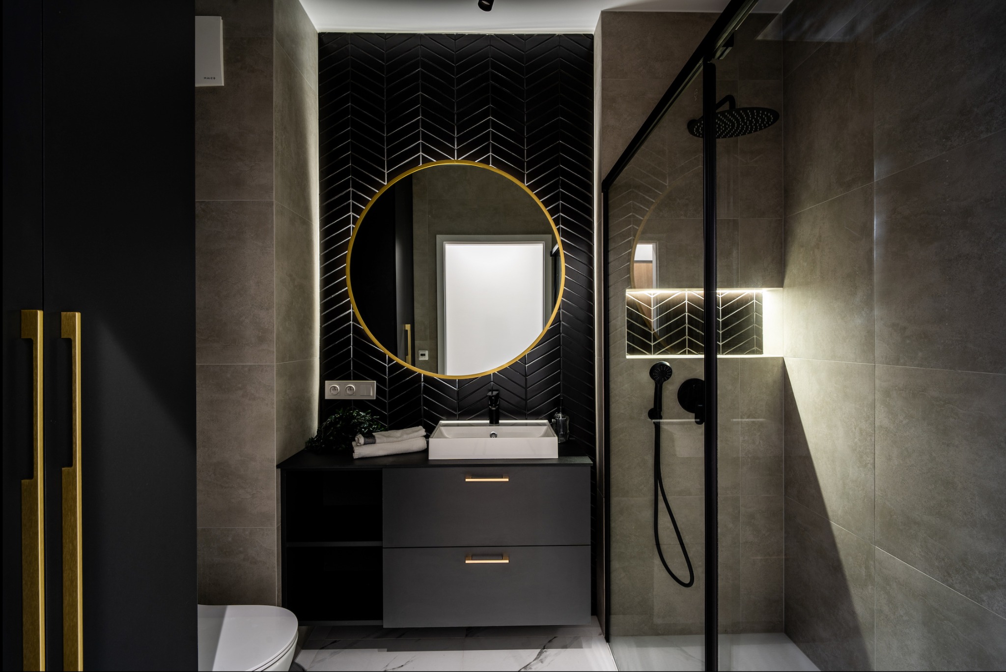 Nowoczesna łazienka z czarną armaturą, okrągłym lustrem w złotej ramie i płytkami ułożonymi w jodełkę na ścianie za umywalką. Kabina prysznicowa ze szklaną ścianą.