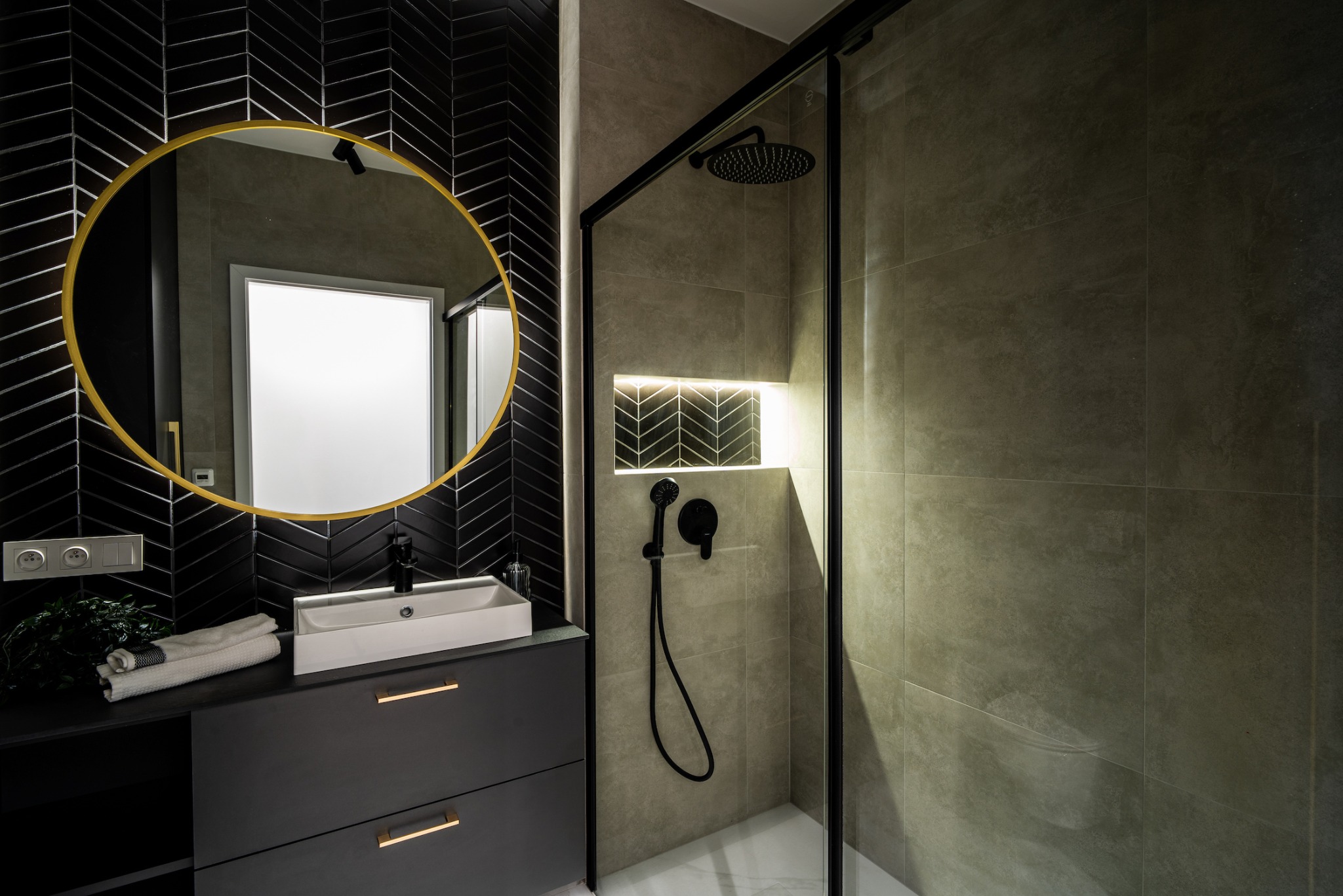 Nowoczesna łazienka z czarnymi płytkami ułożonymi w jodełkę, okrągłym lustrem w złotej ramie, białą umywalką nablatową, szarą szafką z złotymi uchwytami i kabiną prysznicową z czarnymi profilami.