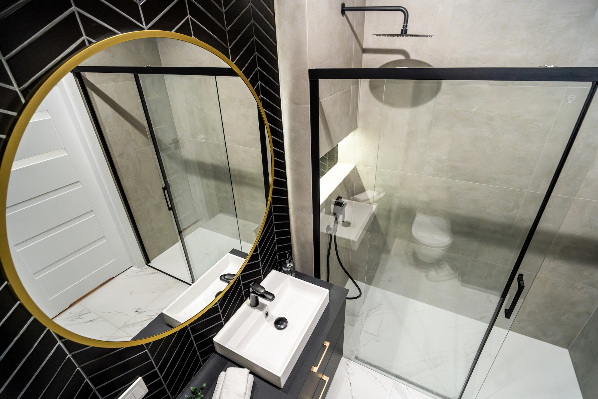 Nowoczesna łazienka z czarno-białymi płytkami ułożonymi w jodełkę, okrągłym lustrem w złotej ramie, przeszkloną kabiną prysznicową z czarnymi ramami i prostokątną umywalką.