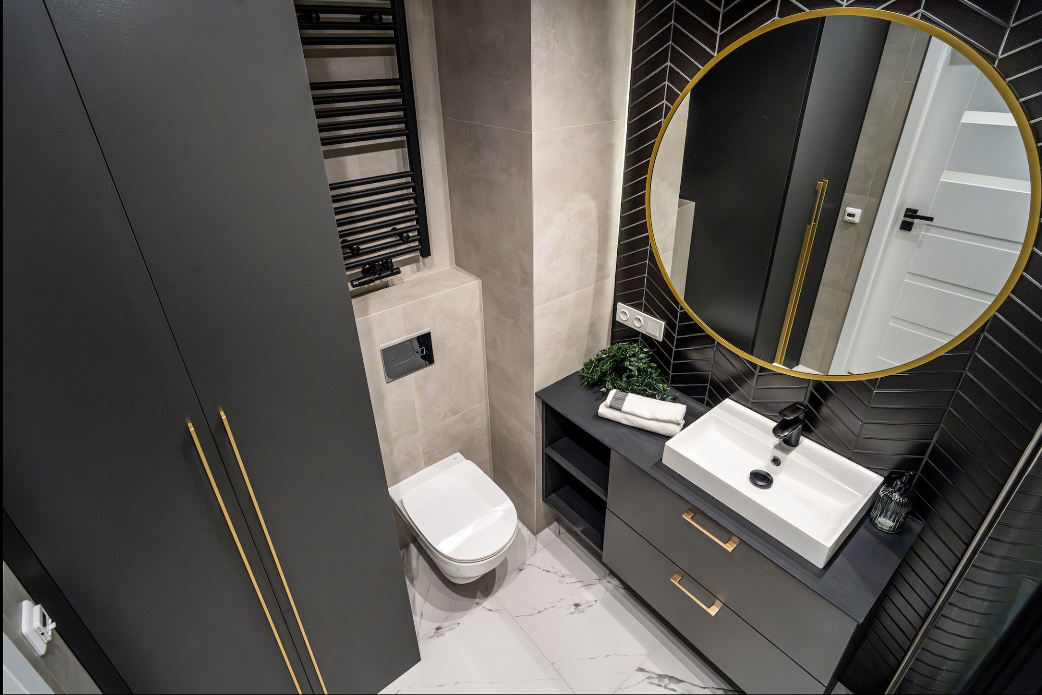 Nowoczesna łazienka z czarnymi szafkami, złotymi uchwytami, okrągłym lustrem w złotej ramie i płytkami ułożonymi w jodełkę na ścianie, widok z góry.