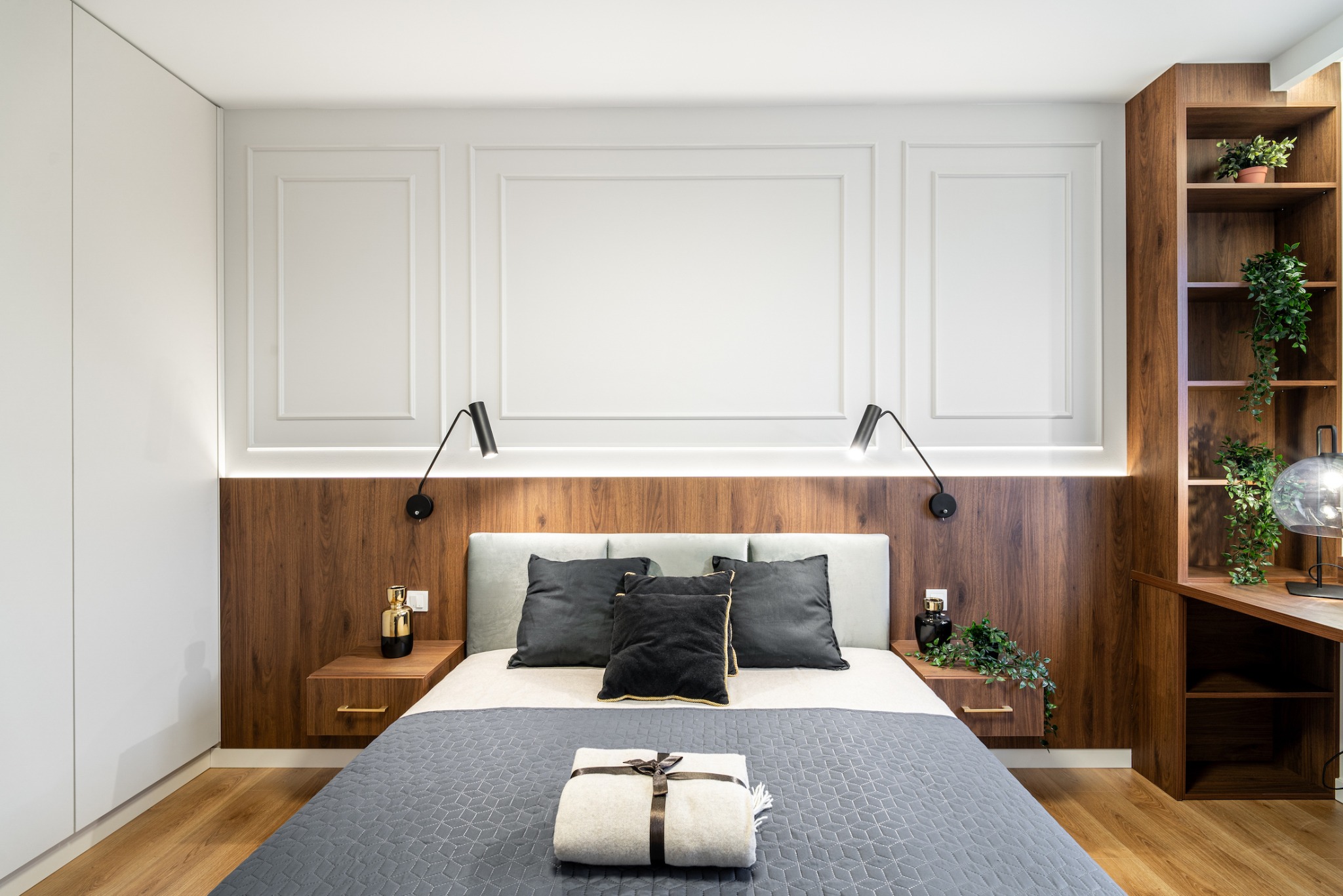Nowoczesna sypialnia z drewnianym wezgłowiem podświetlonym LED, szara pikowana narzuta z ozdobną paczką, czarne lampki nocne i drewniany regał z roślinami.