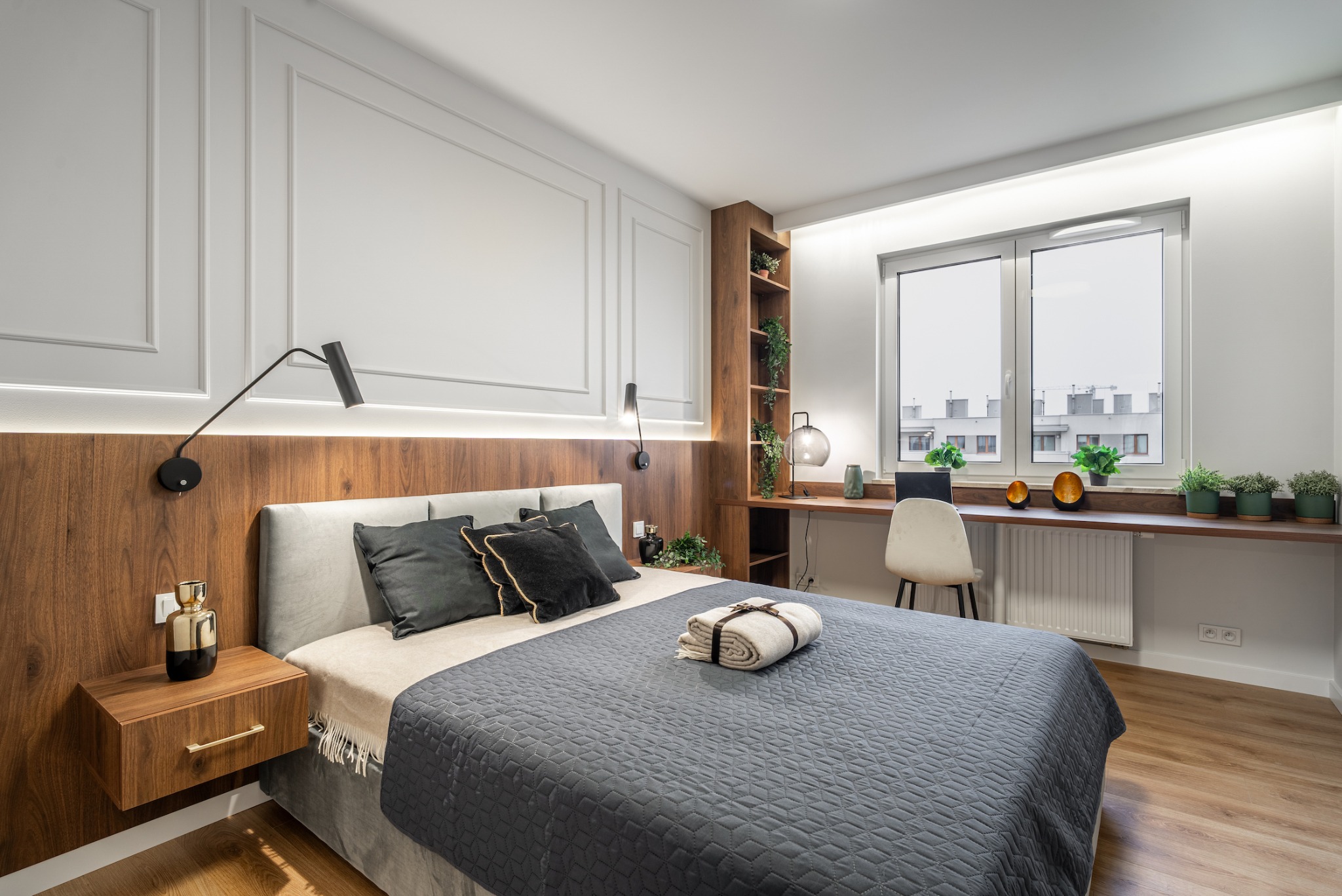 Nowoczesna sypialnia z drewnianym panelem za łóżkiem, minimalistycznymi lampami ściennymi, biurkiem przy oknie z roślinami doniczkowymi i dekoracjami, oraz stolikiem nocnym z ozdobną butelką.