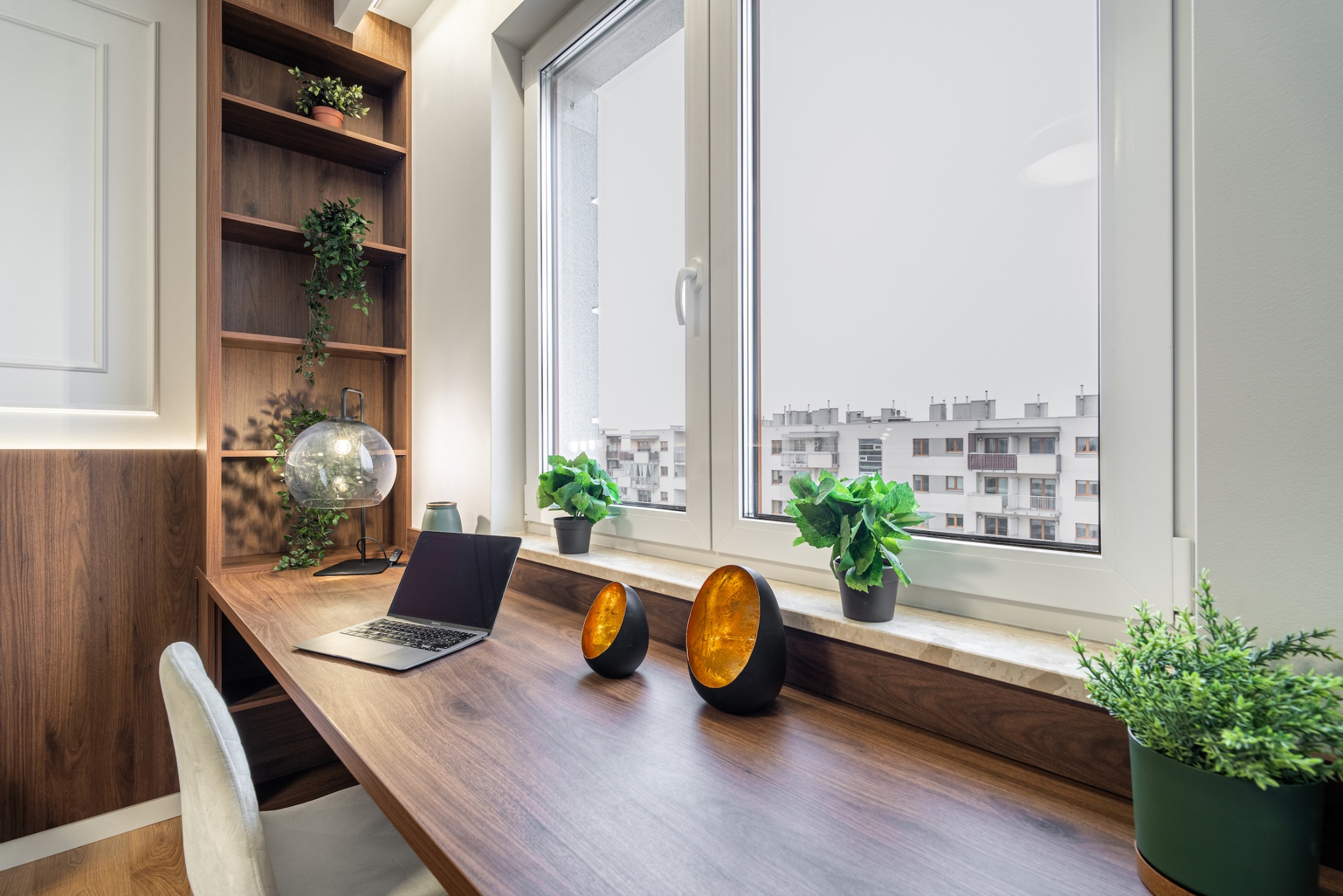 Nowoczesne biuro domowe z drewnianym biurkiem, laptopem, lampą kulą i roślinami doniczkowymi przy oknie z widokiem na miasto.