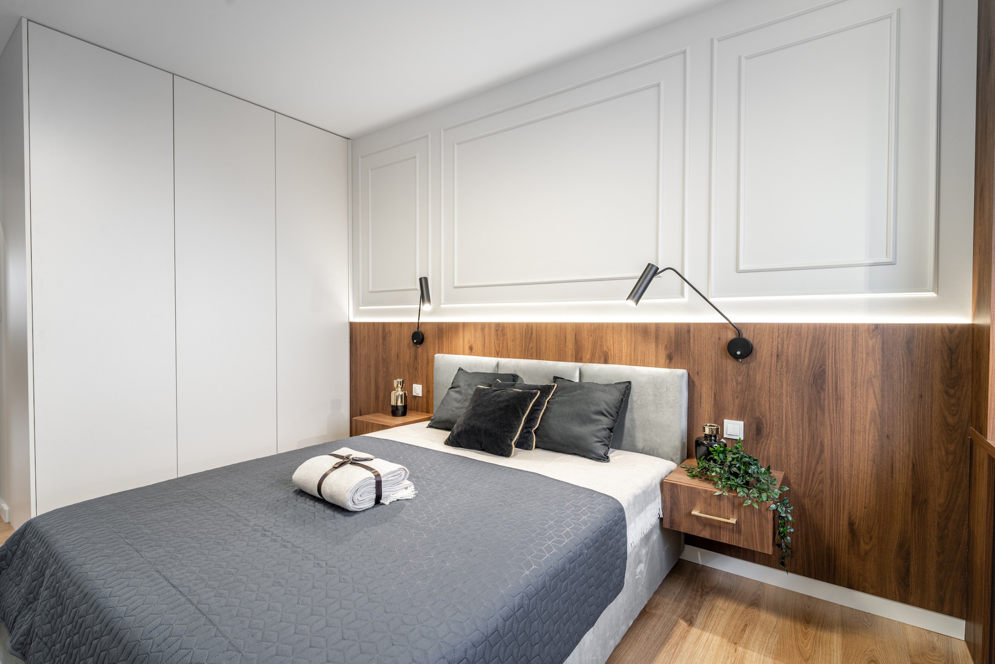 Nowoczesna sypialnia z łóżkiem z szarym zagłówkiem, drewnianym panelem ściennym z oświetleniem LED, białymi szafami w zabudowie i dekoracyjnymi panelami ściennymi, uzupełniona o stoliki nocne...