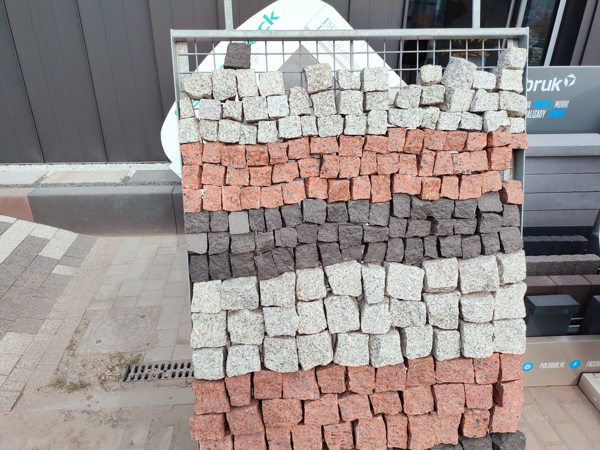 Ekspozycja kostki brukowej w trzech kolorach (szarym, białym i czerwonym) ułożonej w rzędach w metalowej siatce, z widocznym fragmentem chodnika i elementami reklamy w tle.