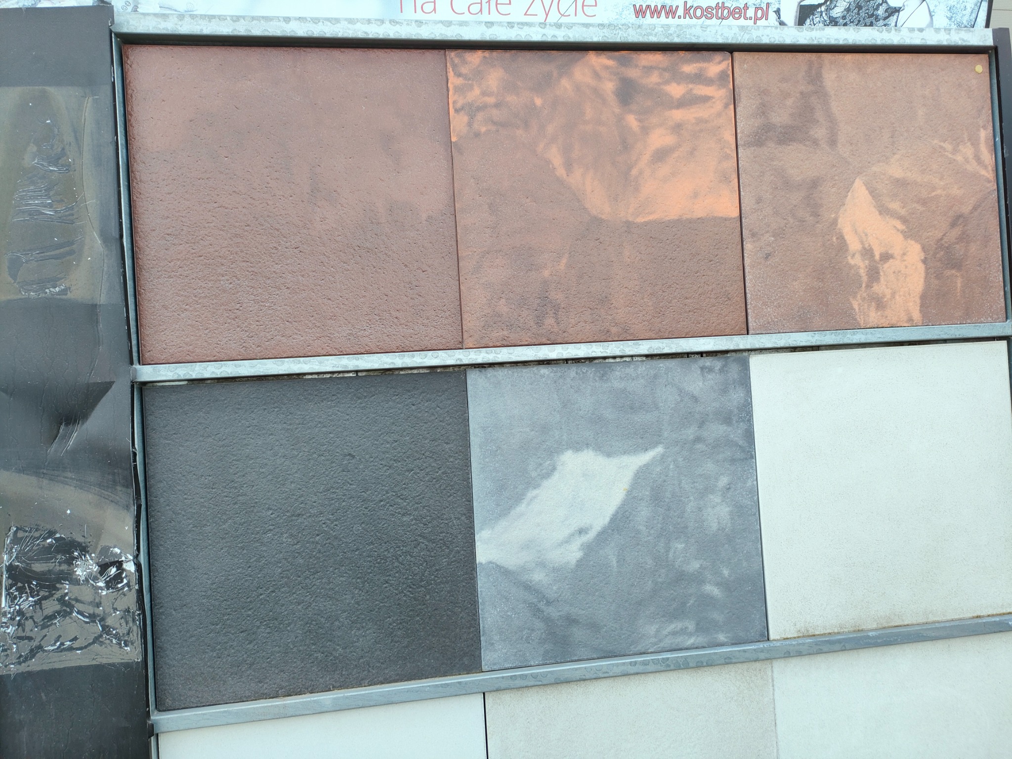 Ekspozycja sześciu kwadratowych płyt chodnikowych o różnych kolorach i fakturach, umieszczonych w metalowej ramie. Górny rząd w odcieniach brązu, dolny w szarościach i bieli. Widoczne logo i adres...