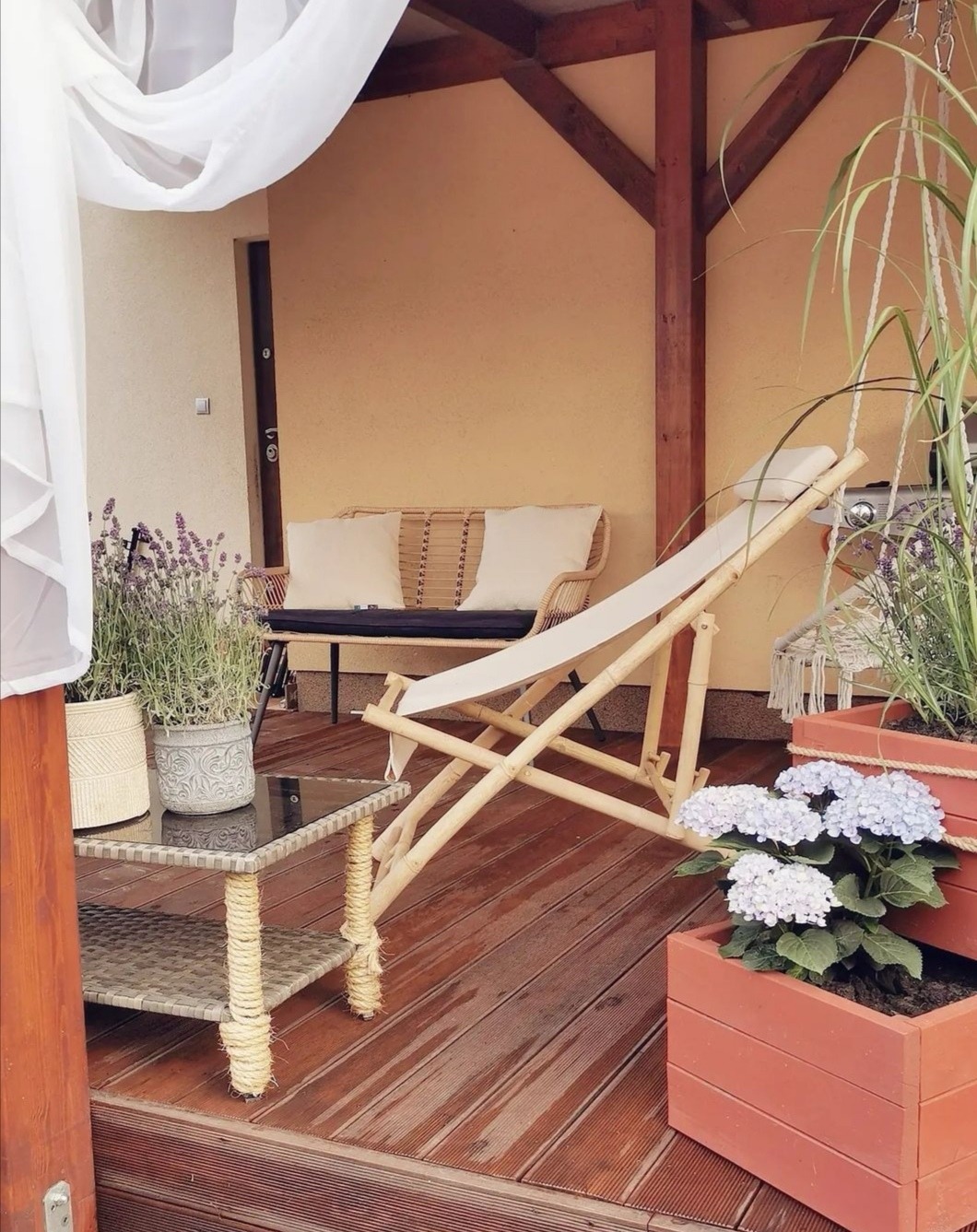 Zadaszony taras z drewnianą podłogą, rattanowym stolikiem i sofą, białym leżakiem, skrzynkami z hortensjami i lawendą, oraz zwiewną białą zasłoną.